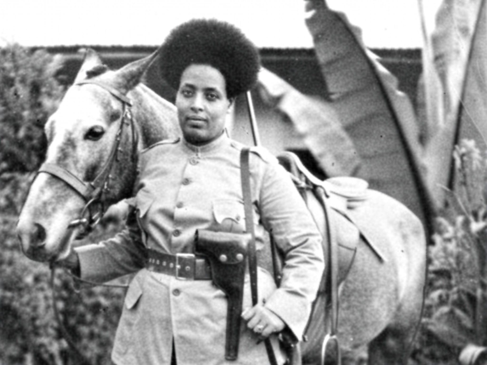 A black-and-white film still from Black Lions – Roman Wolves shows a Black woman with an afro standing forward next to her horse. She is wearing a soldier’s uniform and holding her horse’s reins in her right hand.