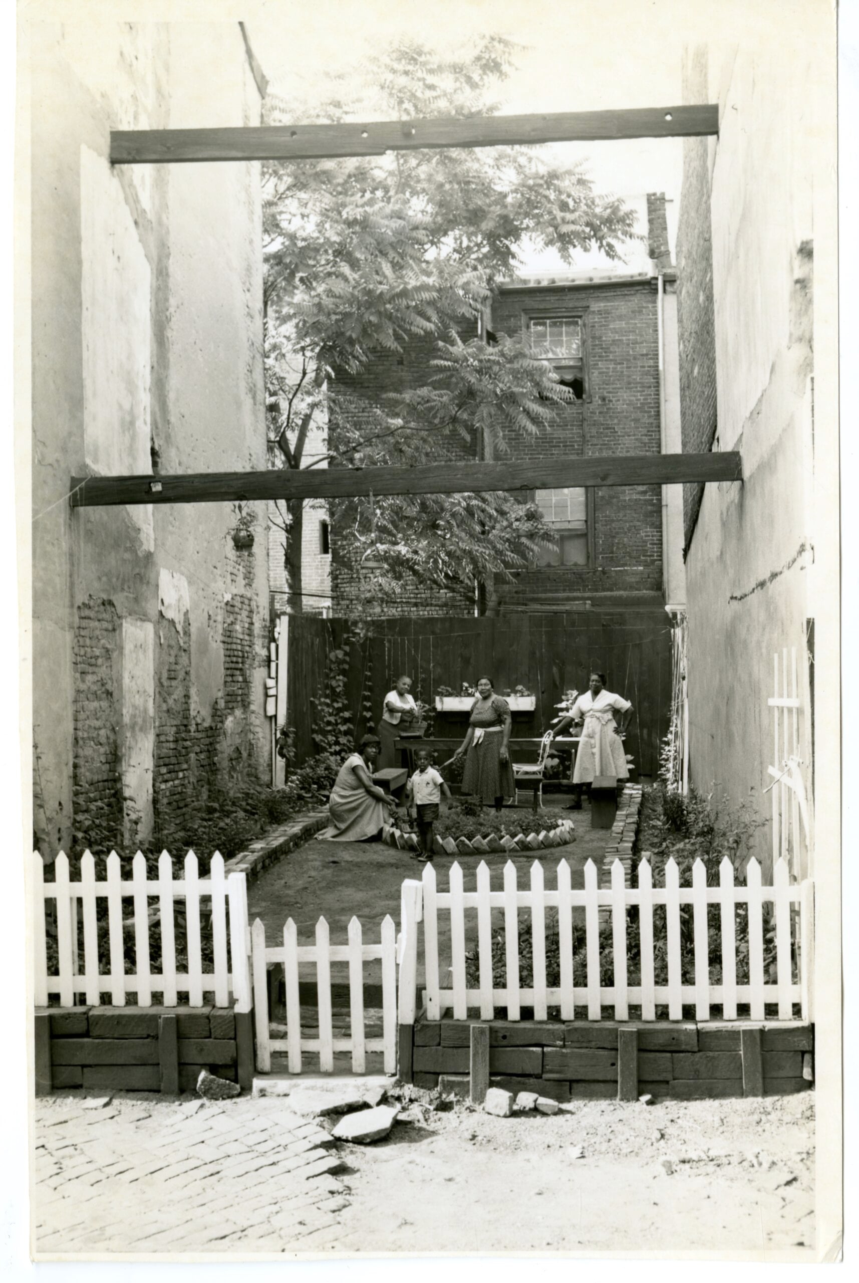 A black-and-white film still from Tenants of Lenapehocking in the Age of Magnets shows a group of four Black women and one Black boy on a lawn. The lawn is surrounded by taller townhomes and blocked from the road with a white picket fence. The group stands around a brick circle with taller foliage.