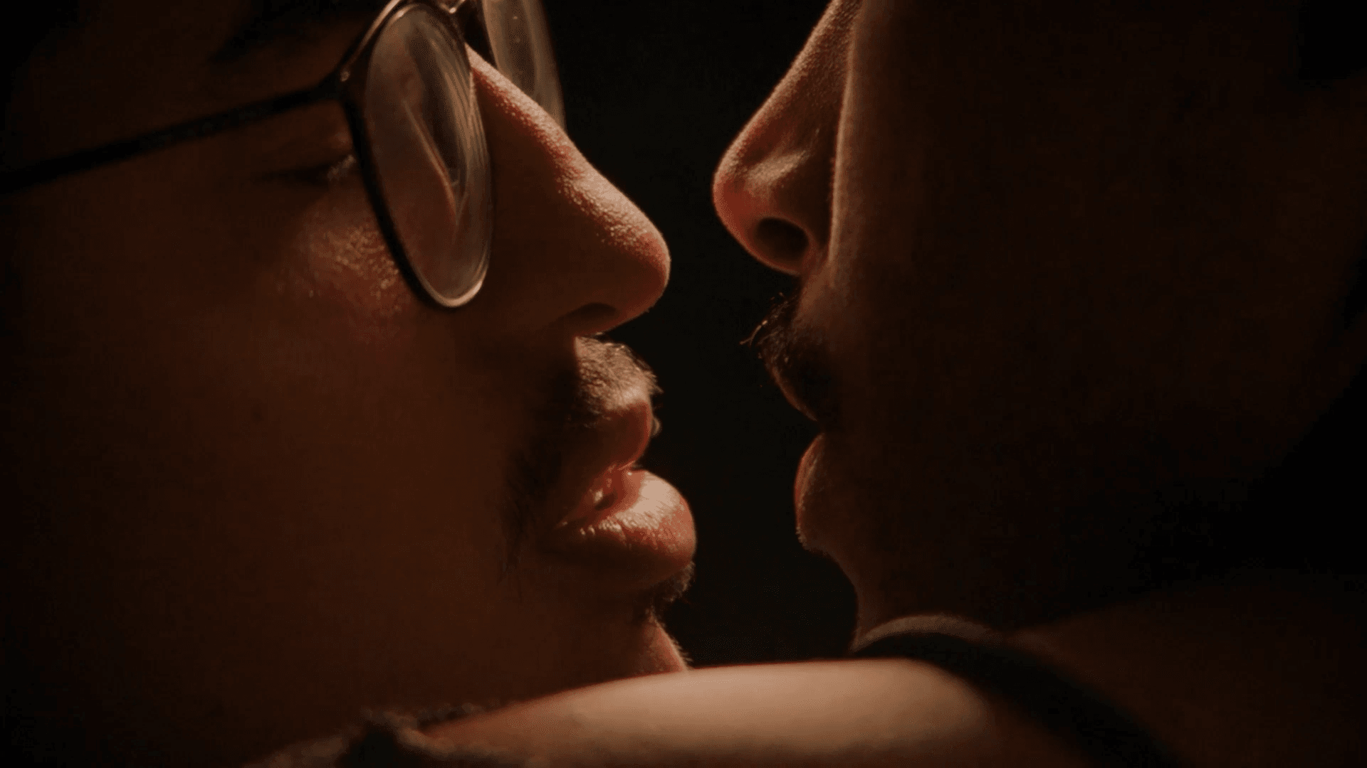 A film still from We Are Not Dreaming shows a close-up of two young Black men with their faces close, about to kiss. The man on the left wears glasses while the man on the right has his arm embracing the other man.