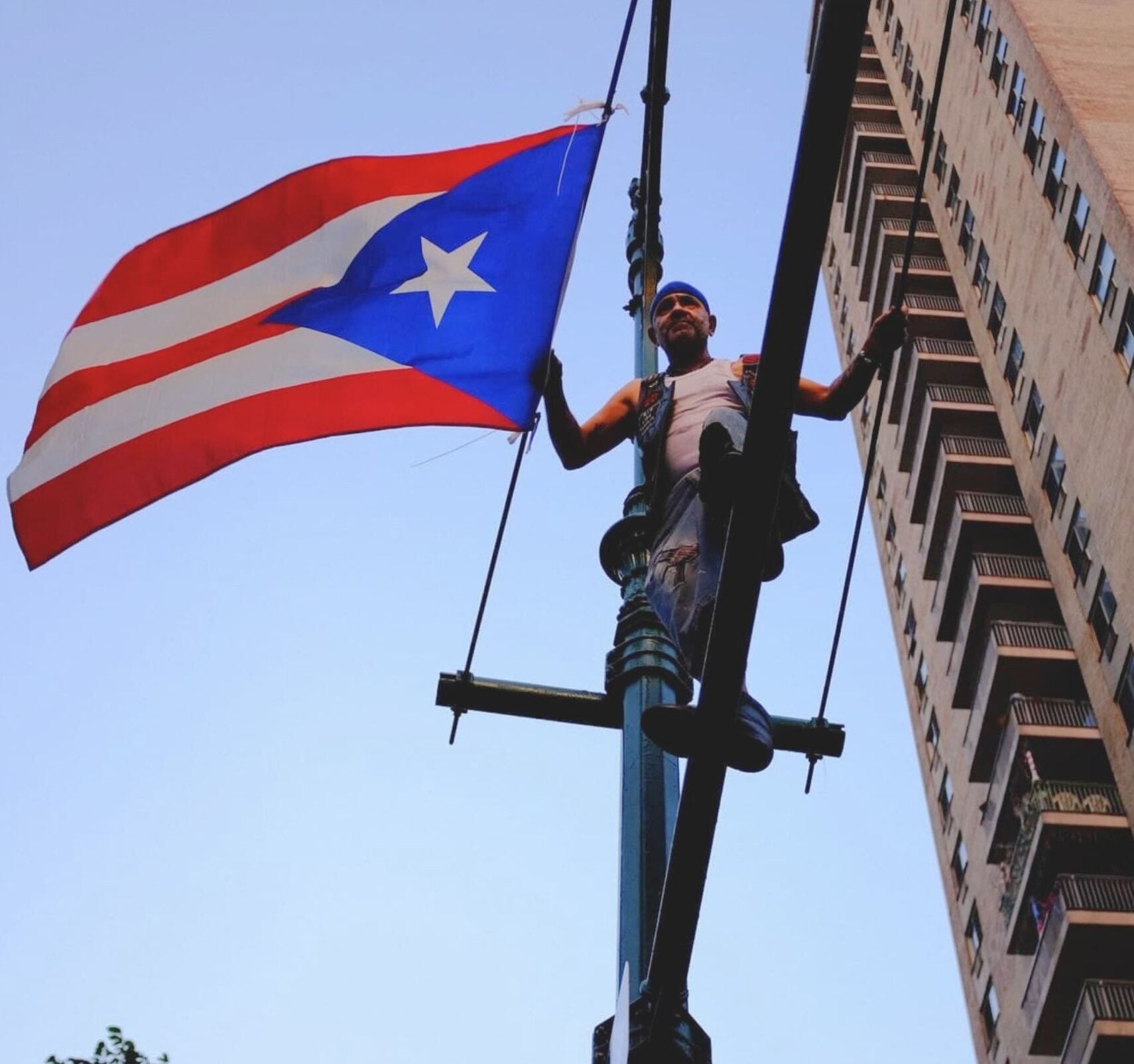 A film still from West Side Familia shows an older Puerto Rican man stands on top of a traffic light pole in New York City, hanging a Puerto Rican flag. The man is wearing a white tank top under a leather vest. Behind him, a tall apartment building stretches into the sky.