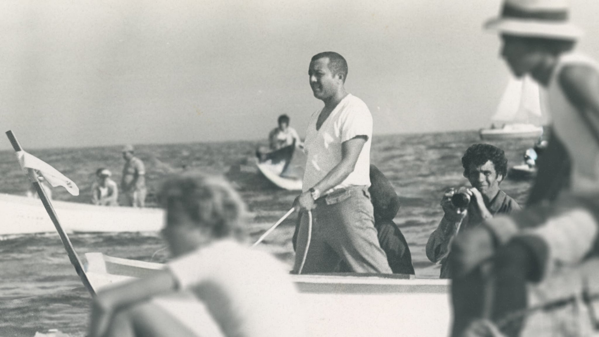 A black-and-white film still from Zenón and the Rebel Boats shows Carlos Zenón standing on a small boat in the Caribbean sea. Zenón is a Puerto Rican man in a white shirt. Around him, there are many other fishermen in boats.