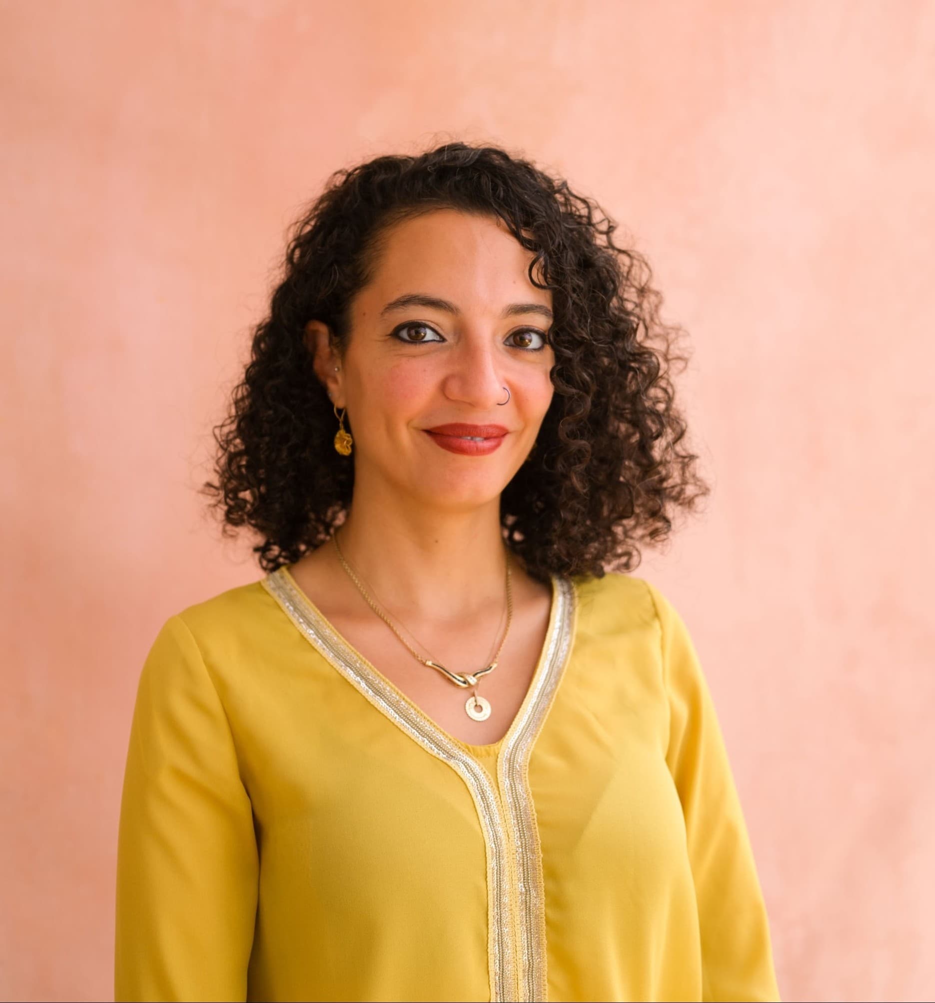 A headshot of Nehad Khader, she is a Palestinian woman. She is wearing a flowy yellow top. She has curly hair. She is smiling softly at the camera.