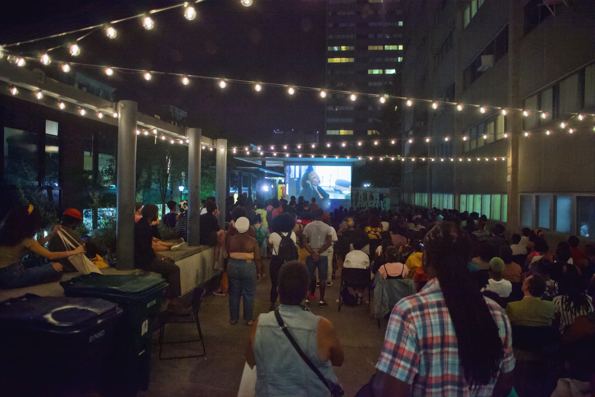 A group of people have their backs turned to the camera as they attend an outdoor film screening, standing under multiple strings of light at night.