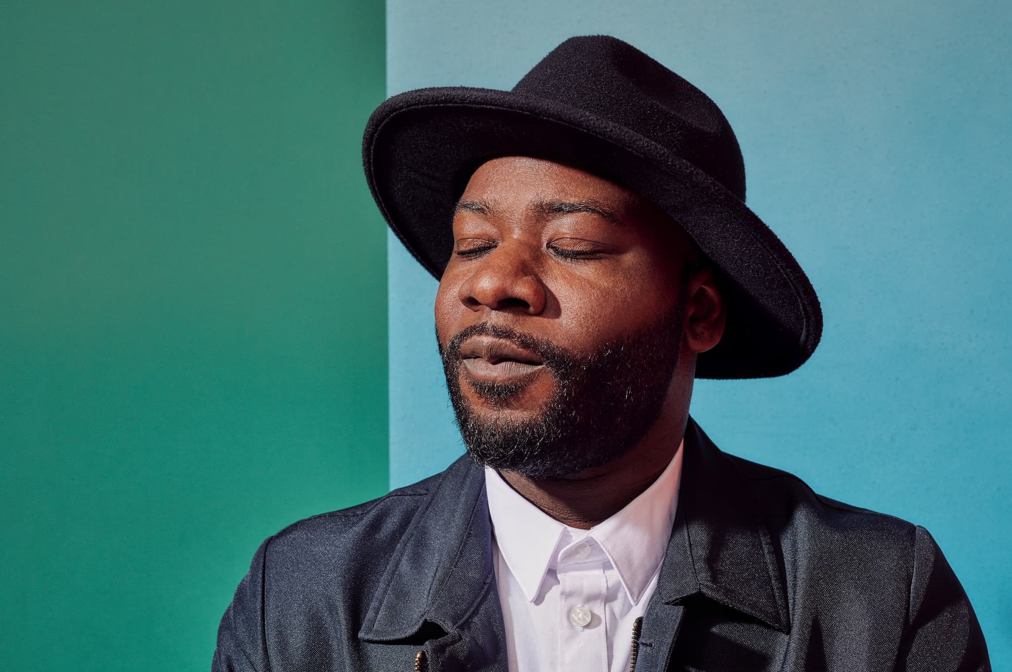 Blitz Bazawule wears a black rimmed hat and a black leather jacket over a white shirt, his eyes are closed as if in meditation. Behind him is a wall which is teal and light blue.