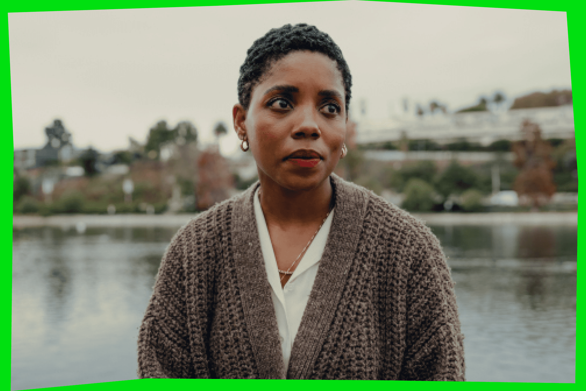 Janicza Bravo photo, wearing a cardigan sweater, standing in front of a body of water, looking off camera to the right. Surrounded by neon green frame.