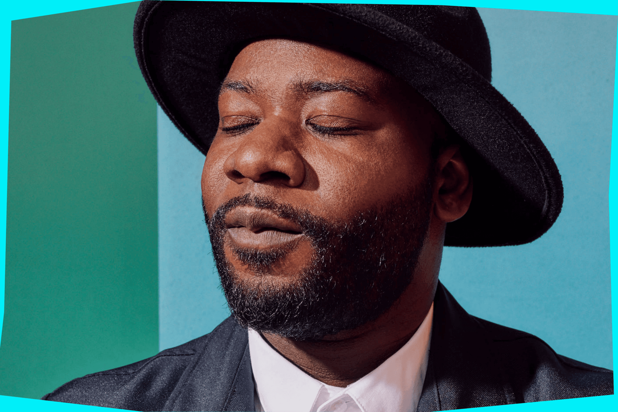Blitz Bazawule wears a black rimmed hat and a black leather jacket over a white shirt, his eyes are closed as if in meditation. Behind him is a wall which is teal and light blue.