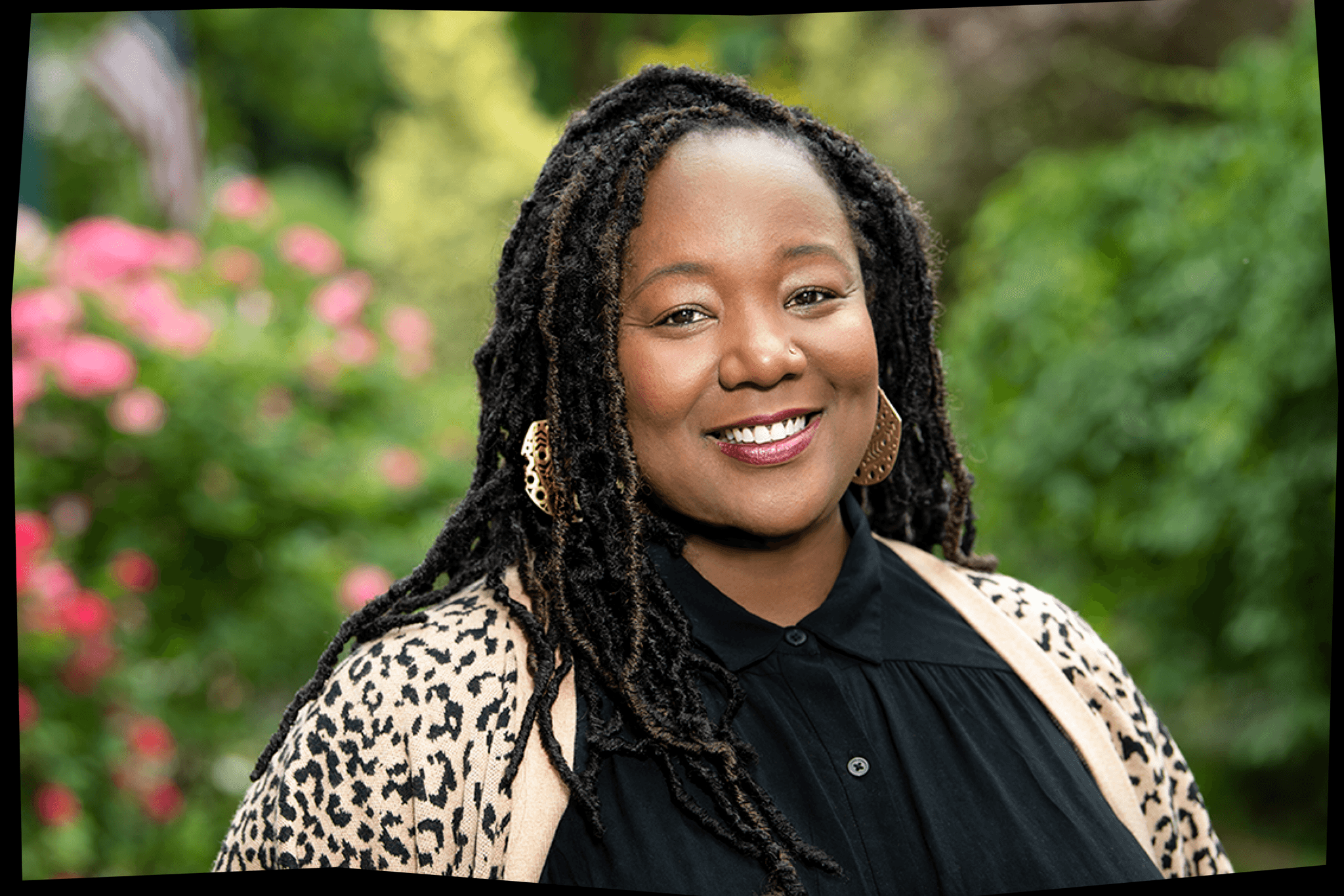 A photo of Maori Karmael Holmes shows her with her hair braided, wearing a leopard print cardigan over a black top, smiling and standing in front of a garden with pink flowers.