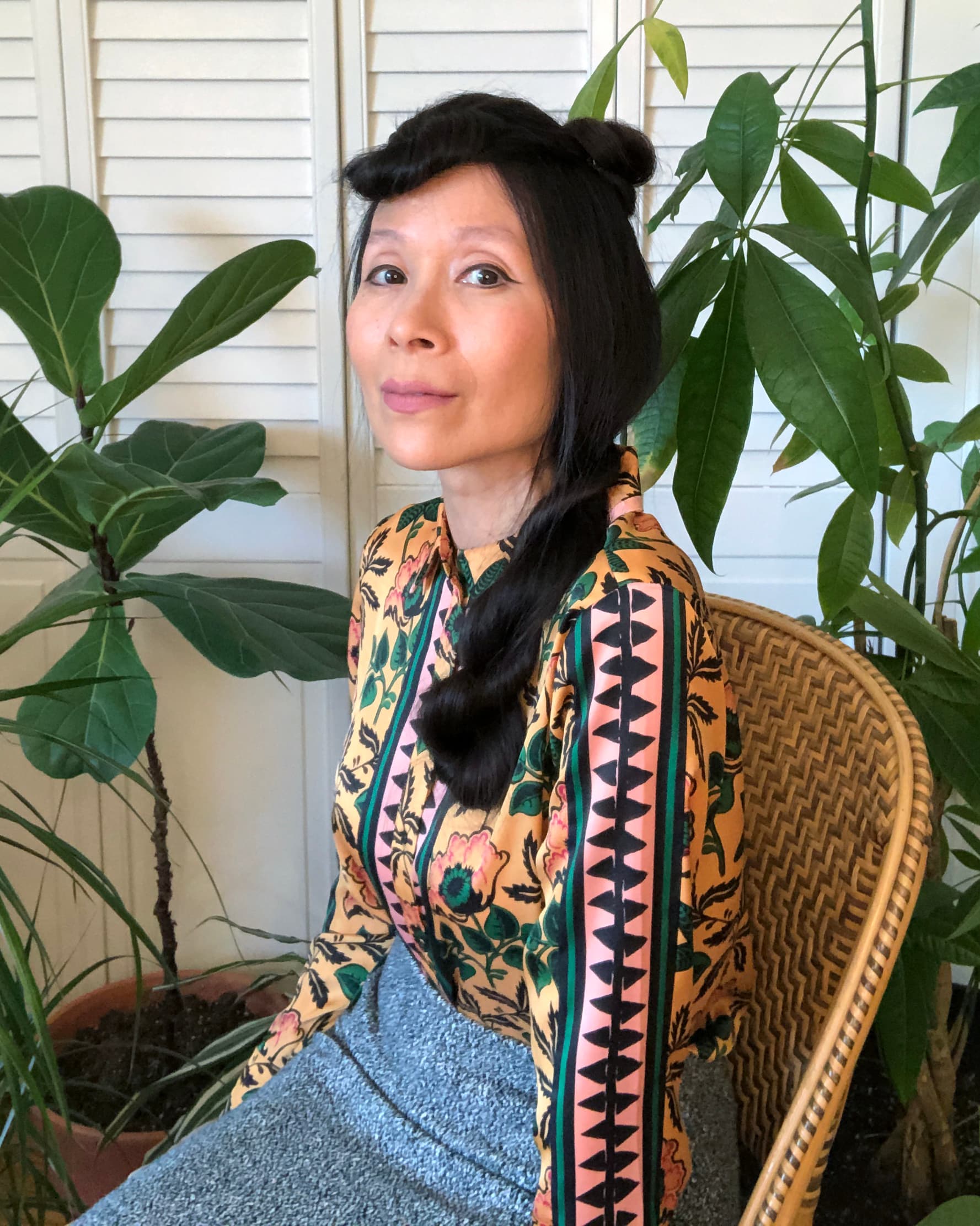 A photo of Astria Suparak. She is sitting in a wicker chair and there are plants behind her. She is wearing a gray skirt and a patterned top. She is smiling and looking at the camera.