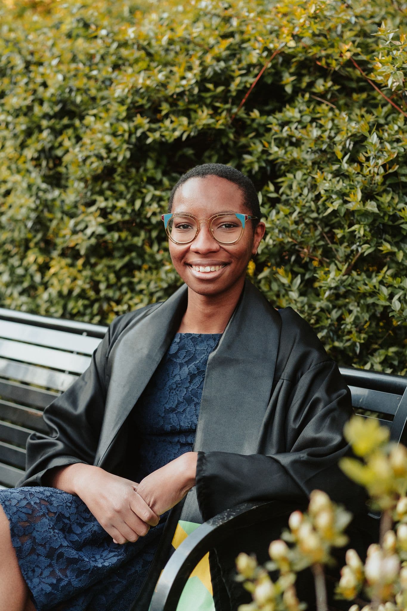 A photo of Jo Brown (she/her/he/him) sitting on a park bench. She wears a black overcoat and dark blue dress. Behind her, green foliage can be seen.