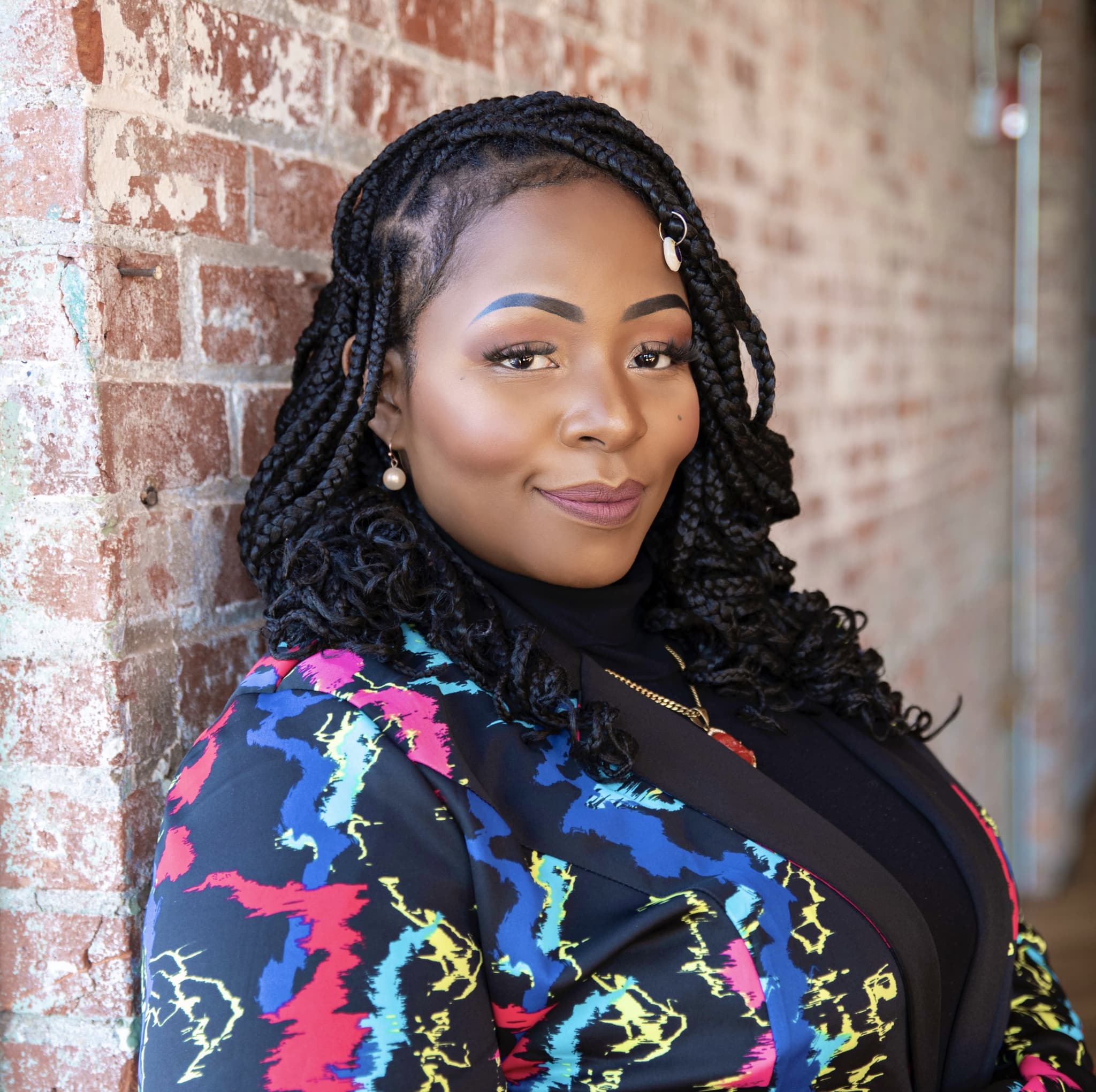 Photo of Sabrina Steward shows her leaning against a brick wall. She wears her hair in long cascading braids and wears a colorful abstract blazer.