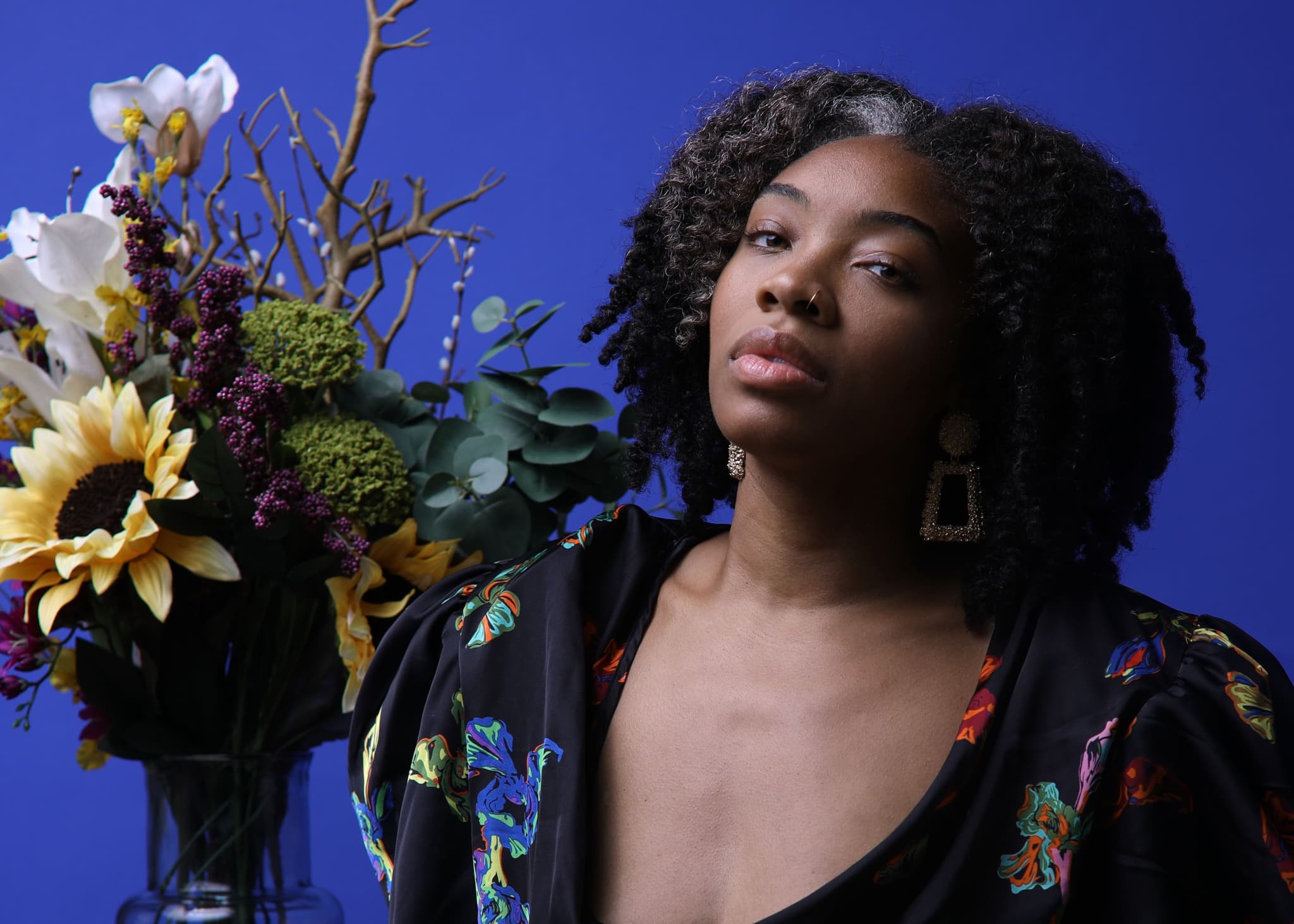 Image of Kerrin Lyons shos her posing with a large floral bouquet filled with various plants and flowers. She wears a black embroidered top with a deep v-neck and tilts her head slightly up as she looks into the camera.