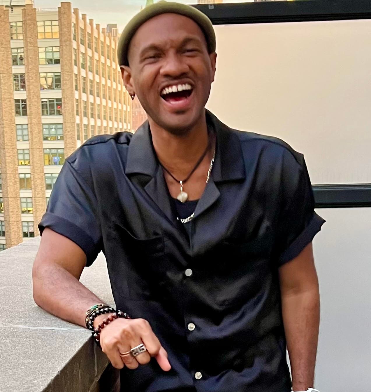 Photo of Michael Moody shows him standing outside on a balcony. He wears a green beanie on his head and a black button-down shirt. He leans forward on his forearm and laughs.