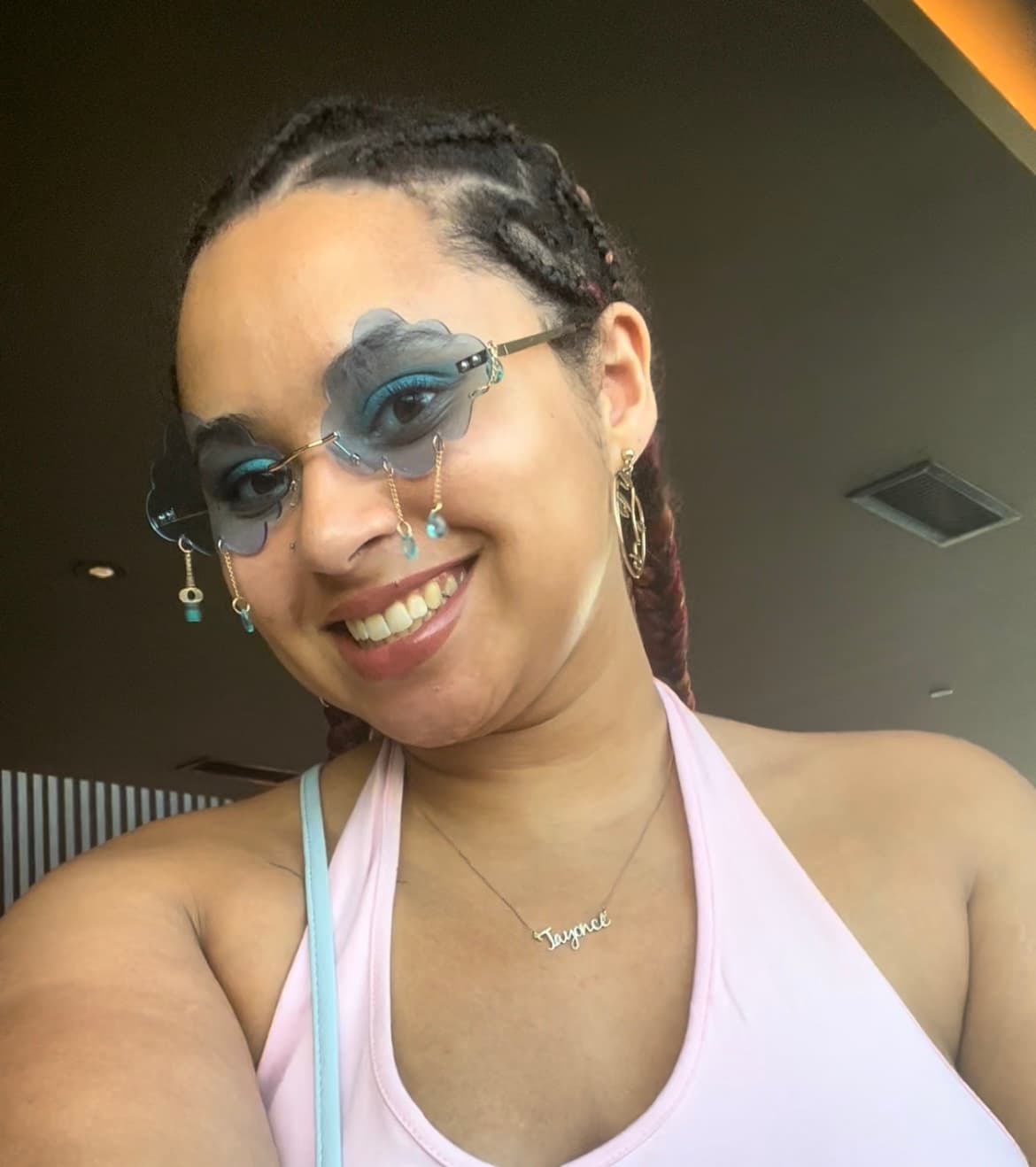 Image of Taylor lamb shows her smiling as she tilts her head to the side. She wears a pink halter top and large statement sunglasses in the shape of blue clouds. Small beads dangle from the glasses like raindrops.
