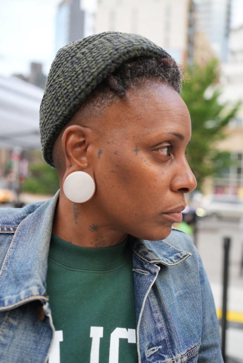 Headshot of the artist Lynneé Denise shows her wearing a jean jacket and green knitted hat. She wears large white ear pluggs in her stretches ears and has several small tattoos of stars and dots scattered on her neck and face.