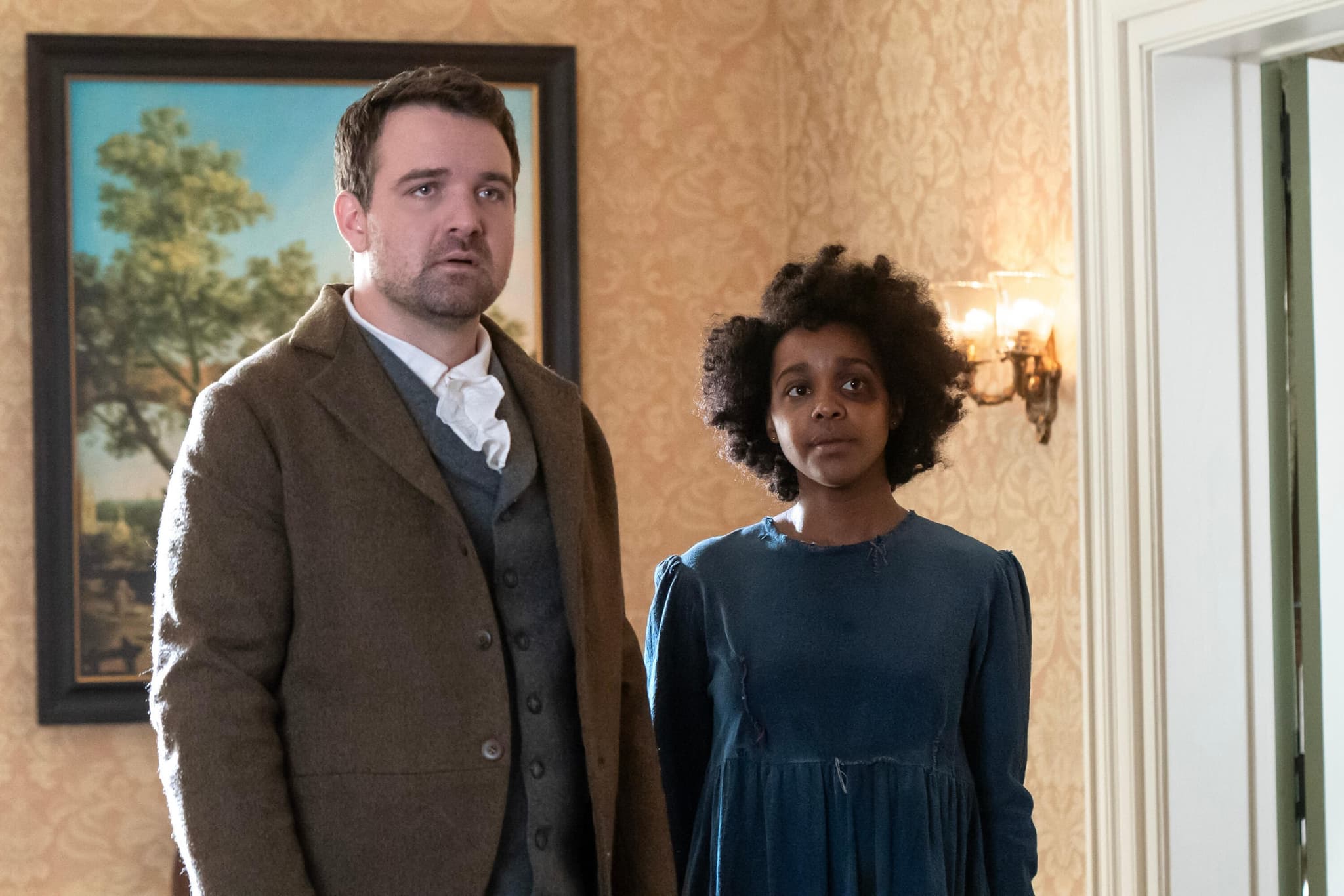 A white man, dressed in a suit, stands next to a Black woman dressed in a worn blue dress, hair frizzy. They both appear to be in a time in the past.