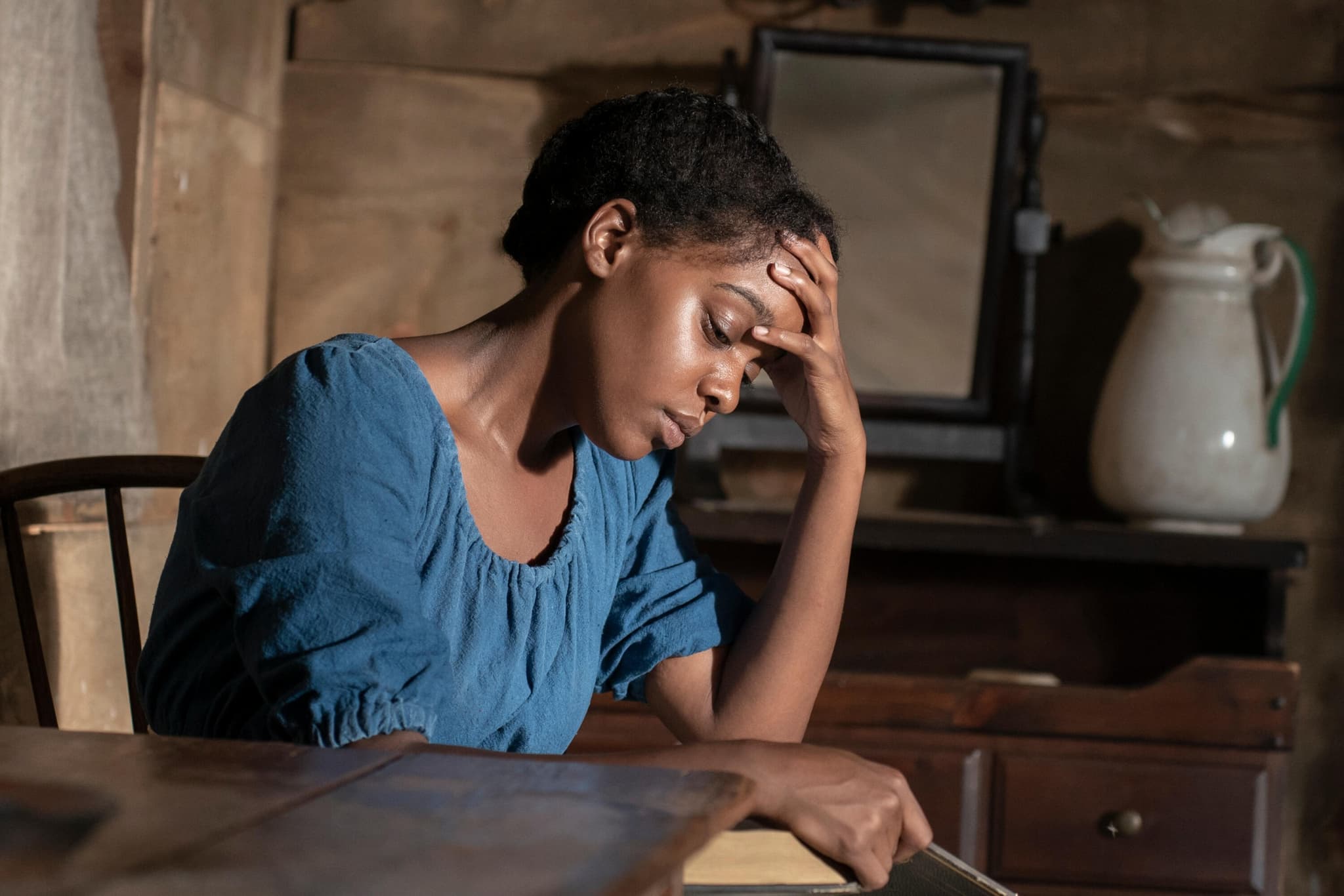 A Black person, the protagonist of Kindred, sits with their head in their hand, dressed in a worn blue dress.
