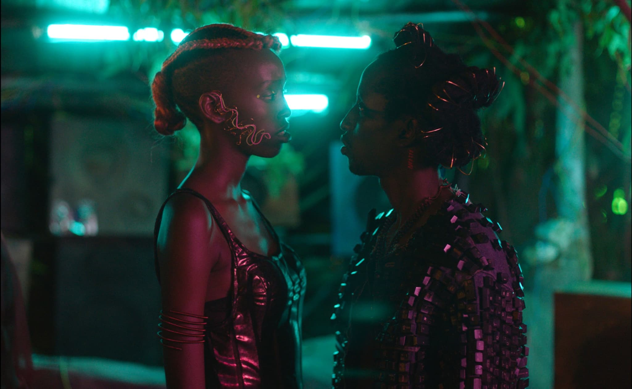A still from Neptune Frost, directed by Saul Williams. Two dark skinned people facing each other in a a techno-fantastic teal lit space.