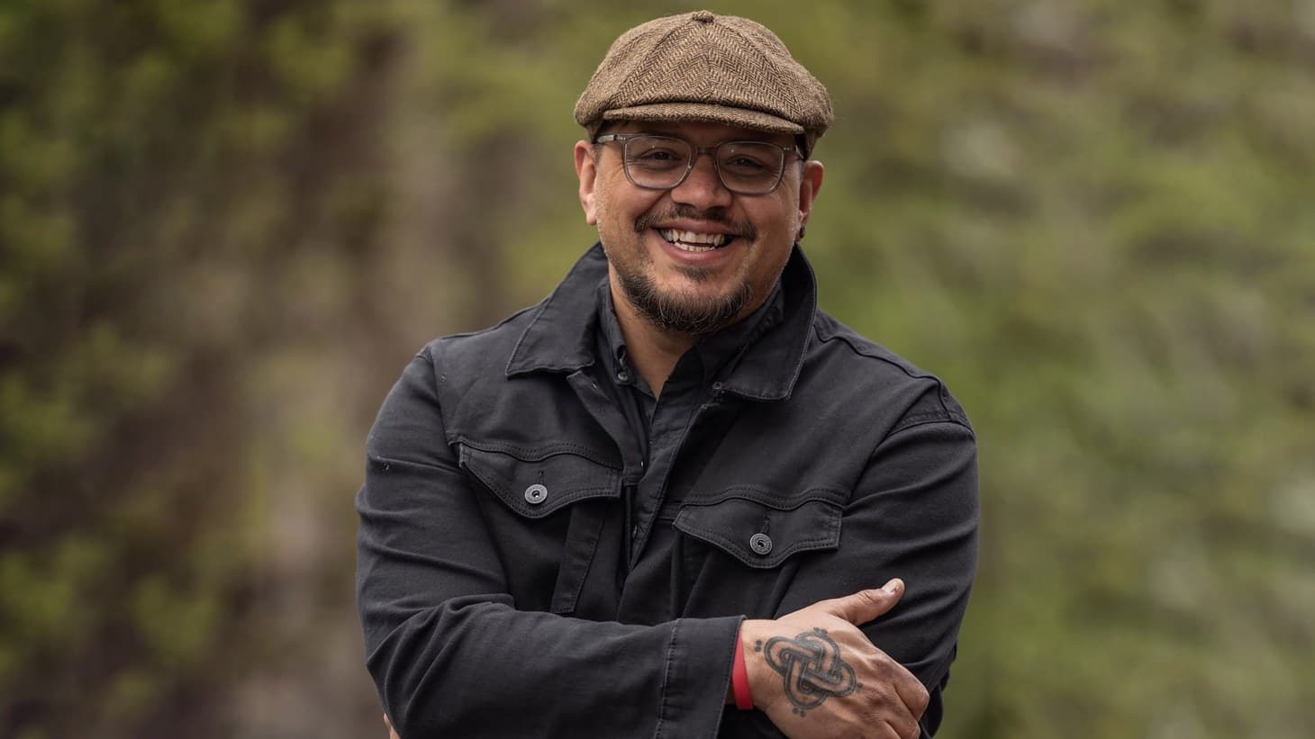 A photo of Sterlin Harjo. He is an Indigenous man. He is standing outside, he is wearing a cap and glasses. He is smiling.