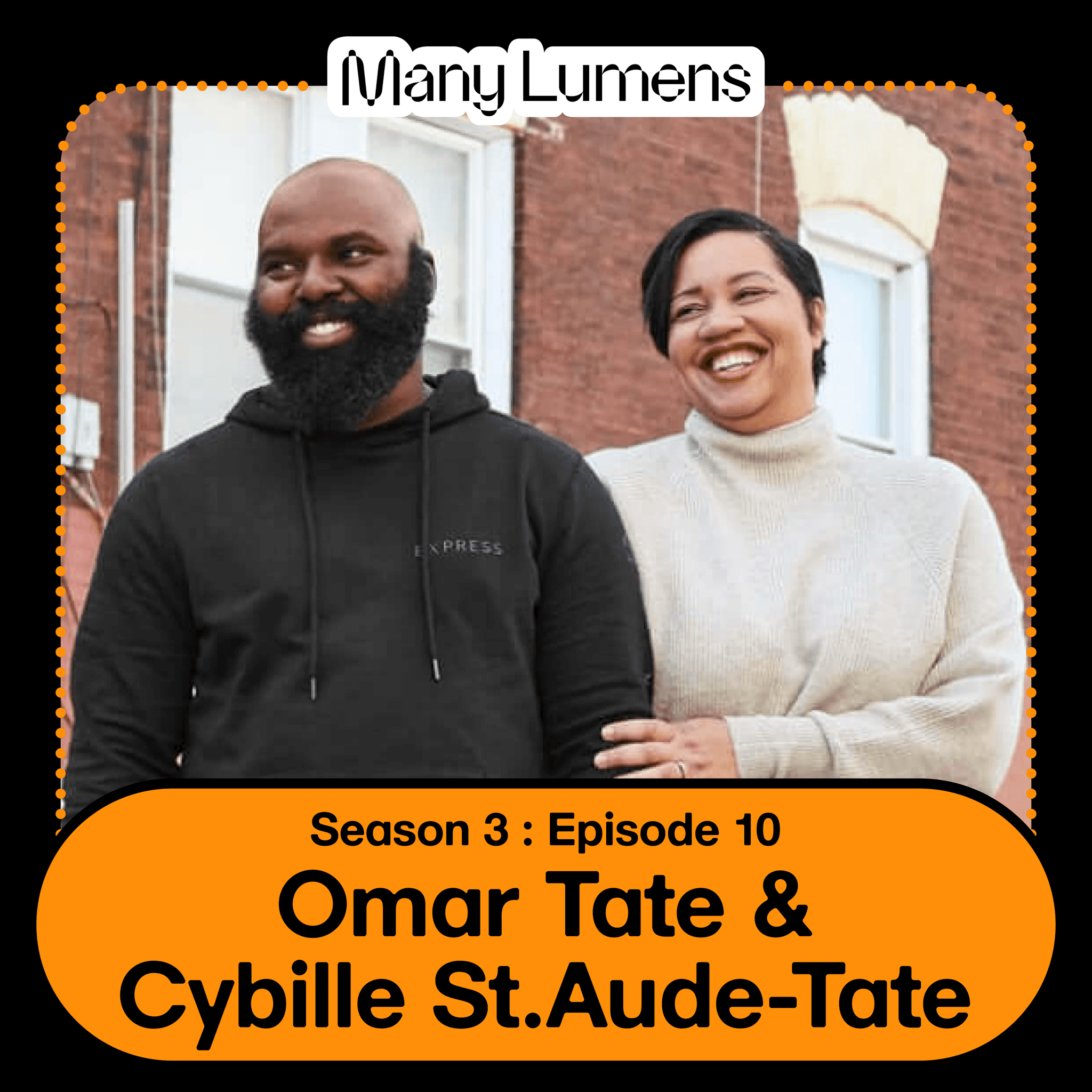 A photo of Omar Tate and Cybille St.Aude Tate. Omar is a Black man, he is bald with a large beard. He is wearing a black sweatshirt and blue jeans. Cybille is a Haitian woman, she has short black hair. She is wearing a white knit sweater and black pants. Omar and Cybille are holding hands and smiling.