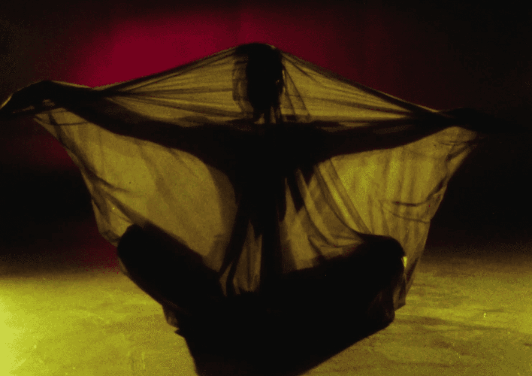 A still from the film Four Women shows a shadowy figure under a blanket, bathed in yellow light and in front of a red wall, only their outline can be made out.