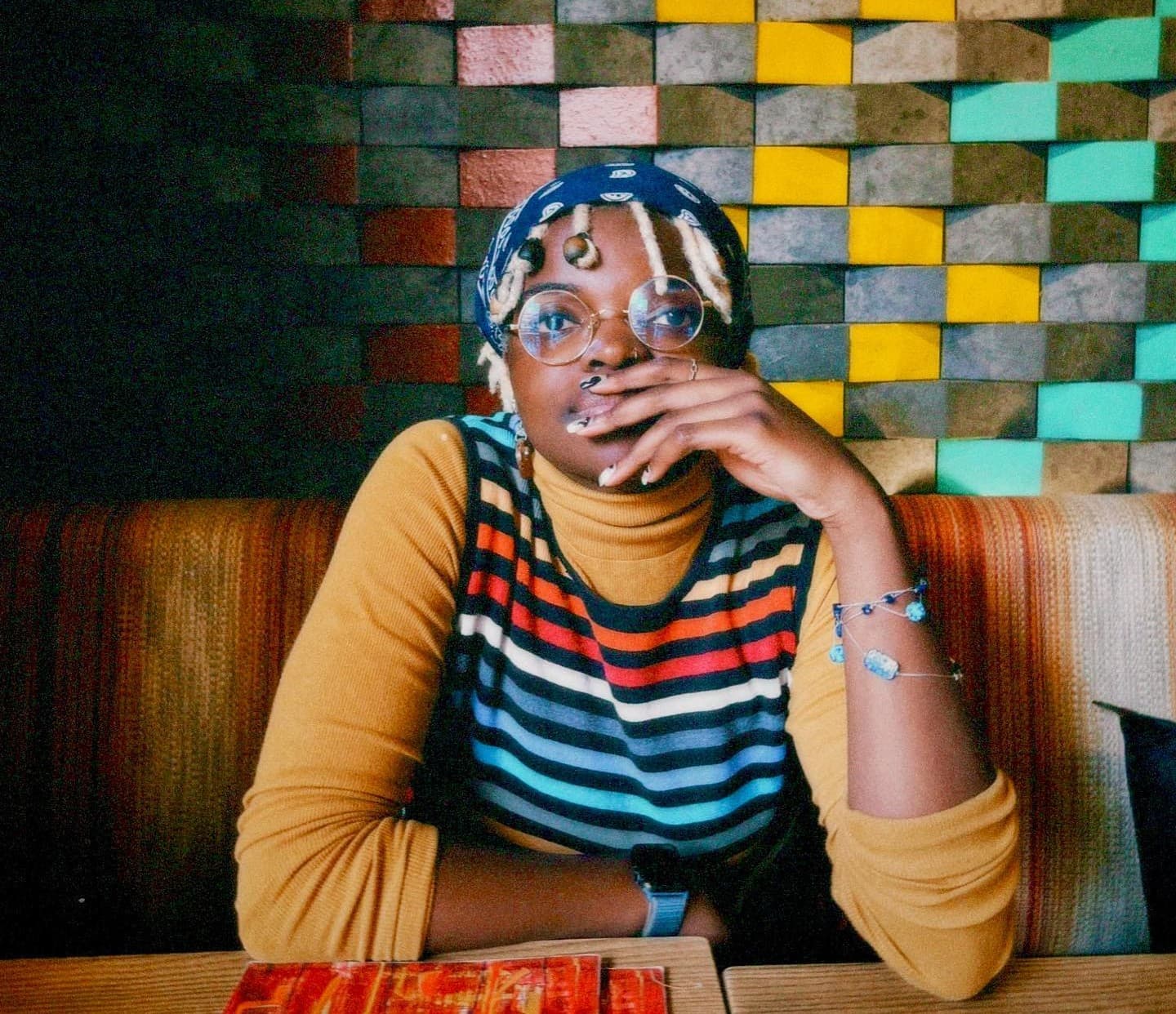 A headshot of Sade, they are a Black person. They are sitting at a booth. The wall behind them is colorful. They are wearing a colorful stripped top and a blue bandana over blond locs.