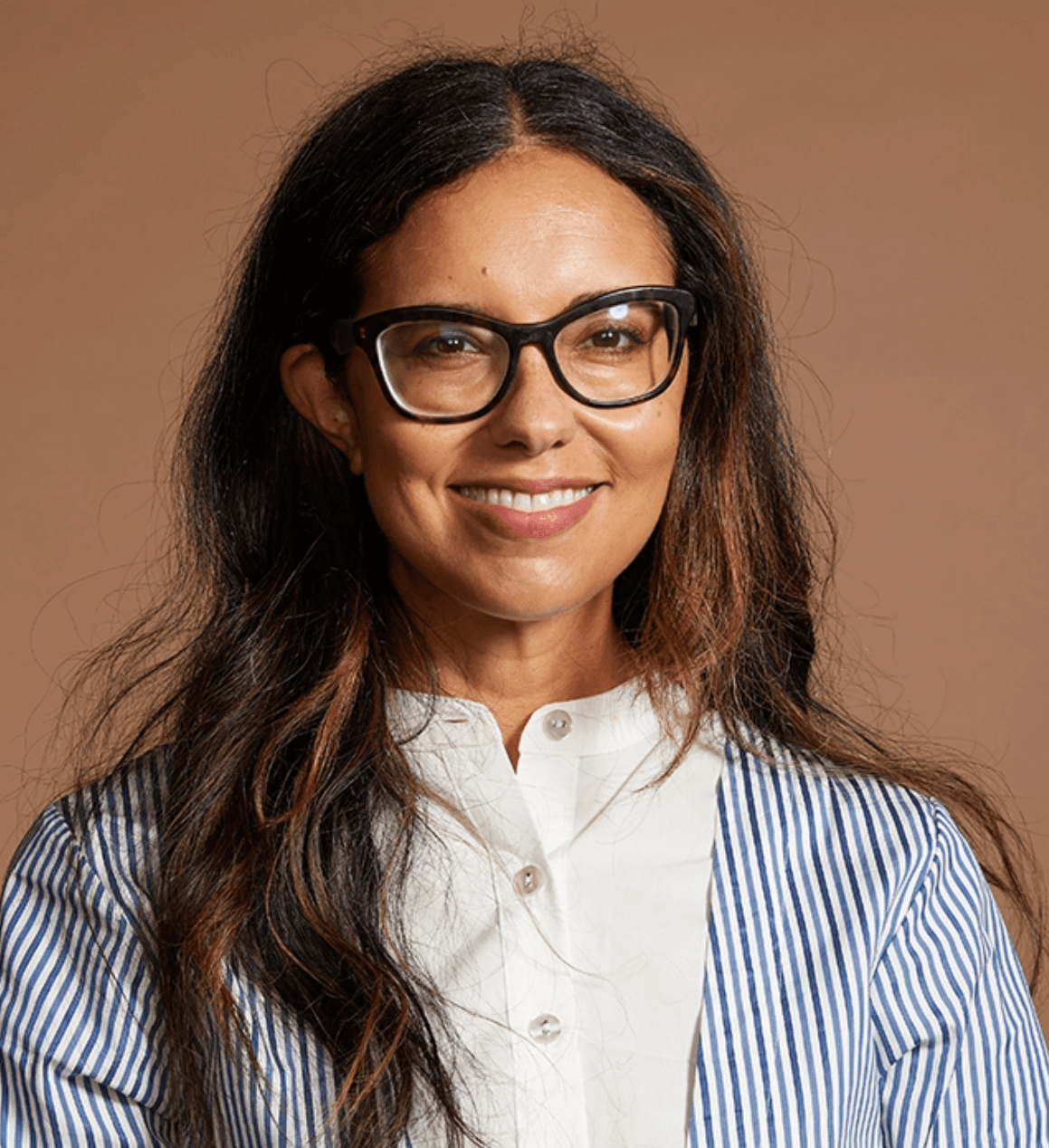 A headshot of Heidi Saman. She is brown woman. She is wearing a button-down shirt, a striped blazer, and glasses. She has long brown hair.