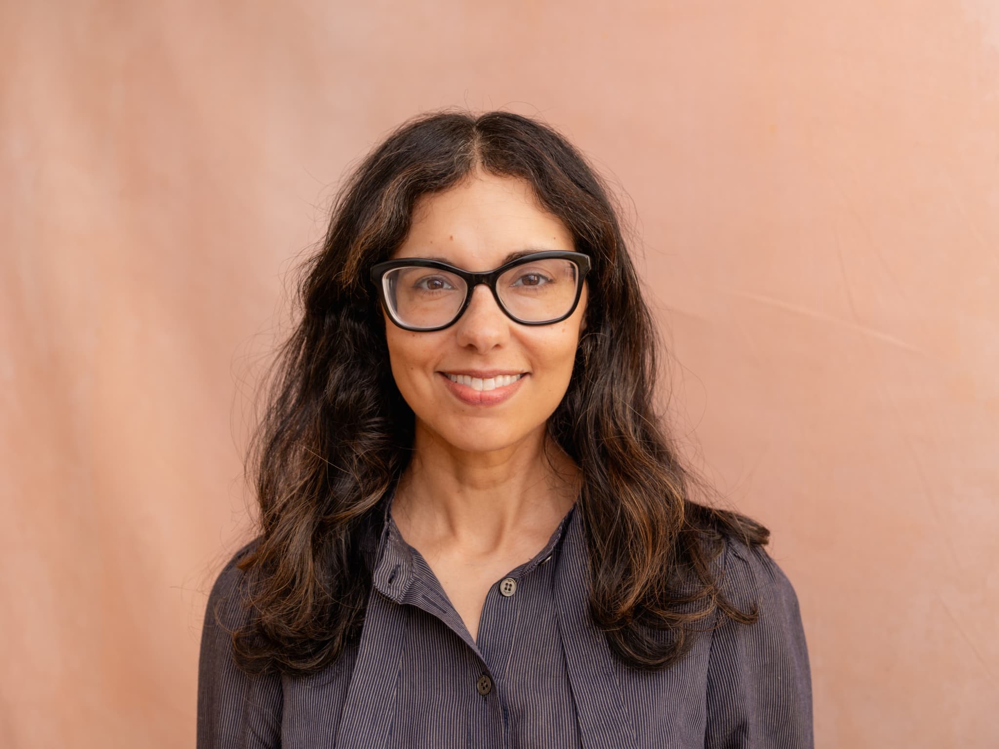 A headshot of Heidi Saman. She is brown woman. She is wearing a button-down shirt and glasses. She has long brown hair.