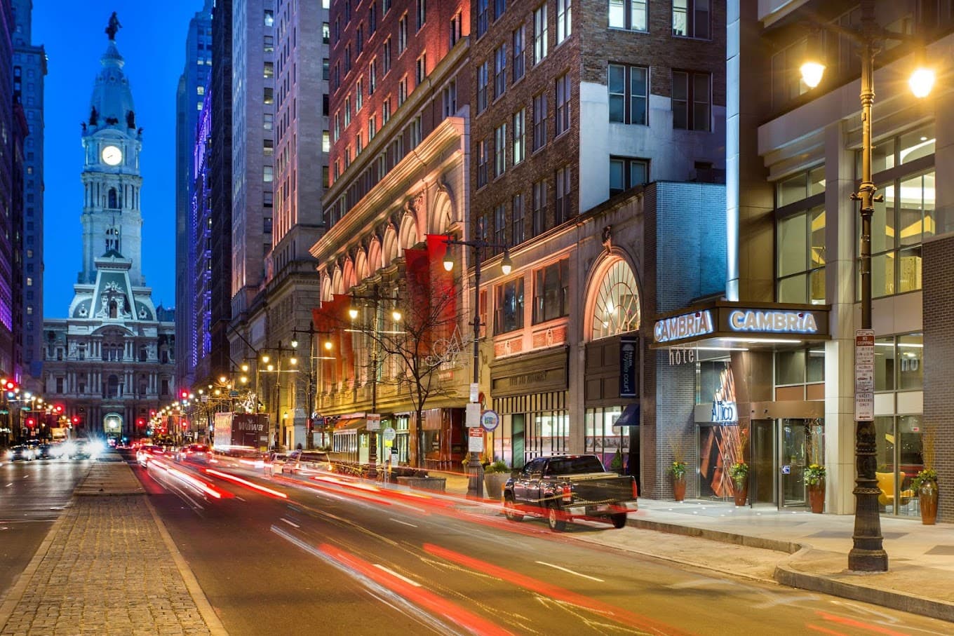 A night time photo of a busy street in downtown Philadelphia, cars zoom by the Cambria hotel, whose awning is prominently visible.