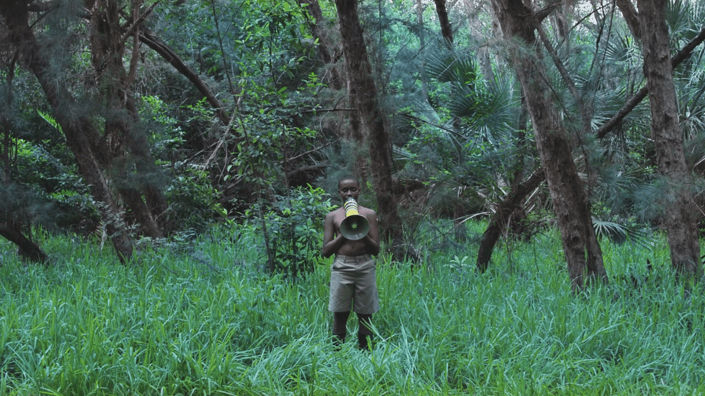 A film still shows a Black person with a bullhorn standing in a green field within a forest