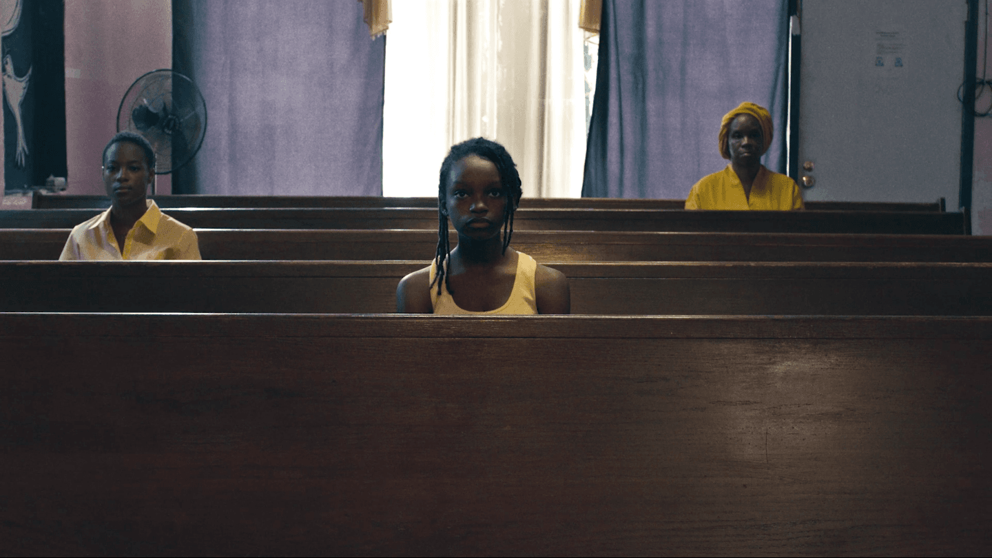 A film still shows three Black people sitting in three different church pews, the one in the center is a child and all wear yellow clothing.
