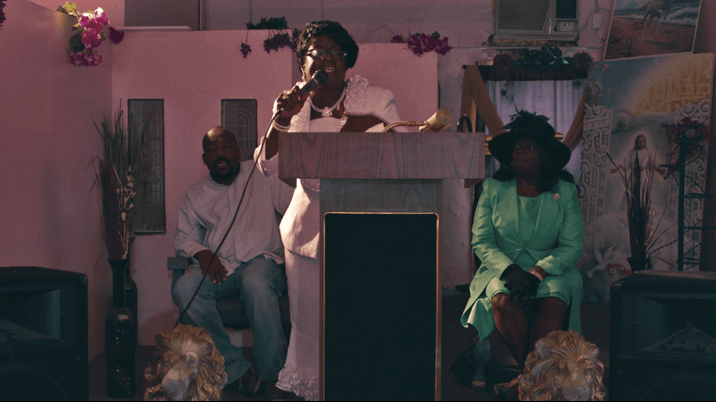 a film still shows a Black woman speaking into a microphine inside a church, presumably giving a lecture.