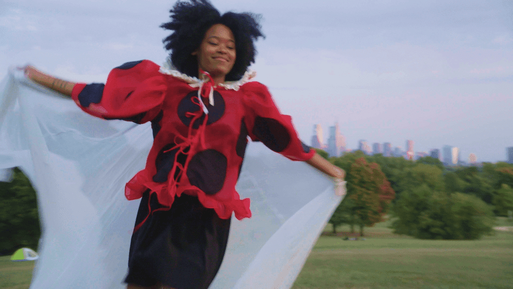 A still from Ladybug shows a young Black woman running through a grassy field. She is wearing a red and black polka-dotted shirt and a white sheer cape.