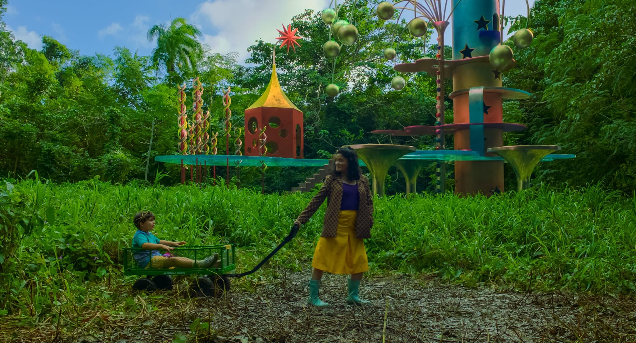 A still image from “Problemista” (2023) showing a young boy in a wagon being pulled by a young woman in a yellow skirt and green rain boots through a grassy area with a kids playground in the background.