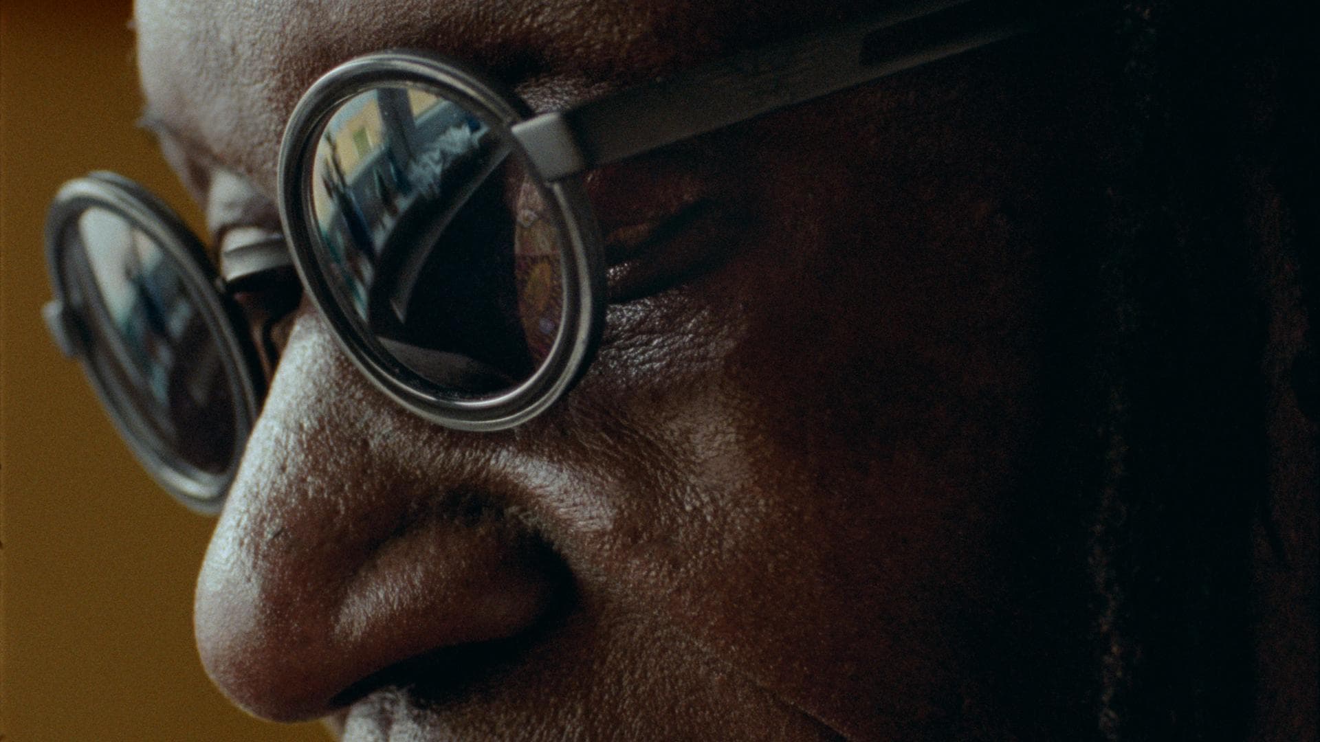 A film still from A LUTA CONTINUA // ATARAXY 44 shows the eyes and nose of an older Black man who is staring at something off screen. He is wearing black circle glasses with dark lenses. In the reflection of the lenses there is a window with items placed along the window sill.