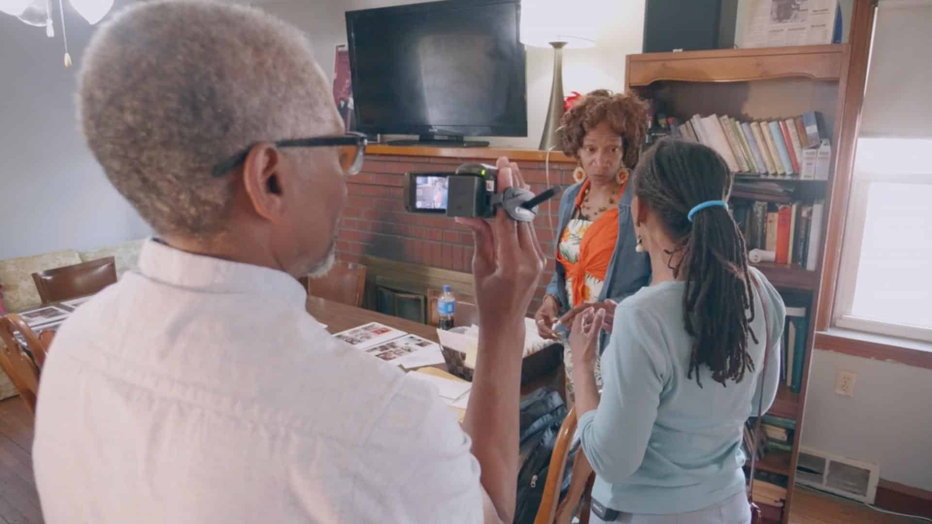 A film still from A New Voice shows two Black women in conversation presumably in a dining room with wooden furniture. The older woman is wearing an orange floral dress and gold and orange pendant earrings. The other woman with her back to the camera has locs tied together with a blue hair band. There is an older Black man recording the women with a camcorder.