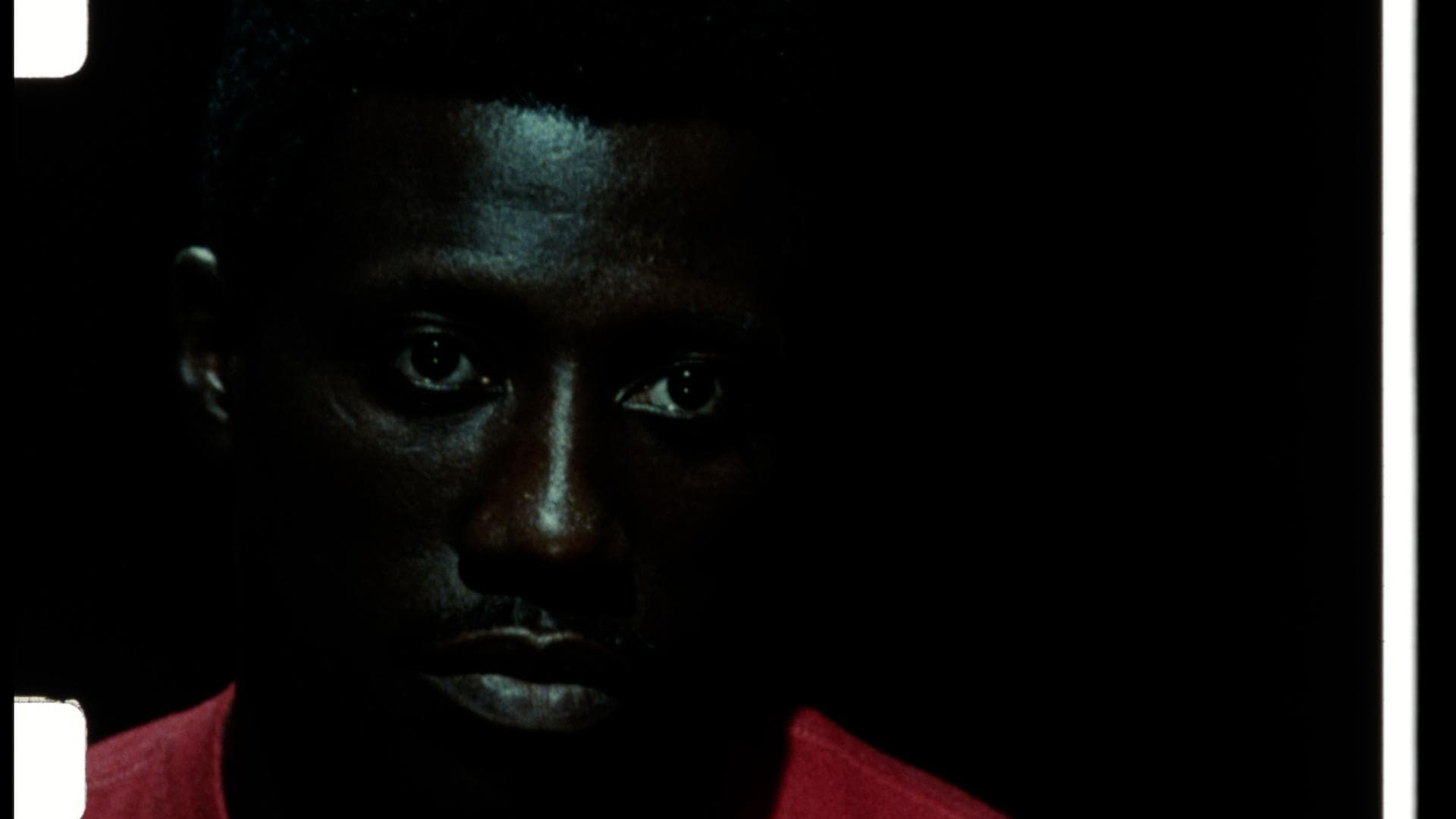 A film still from Another One shows a Black man in a dark room. There are highlights only on his forehead, nose, and mouth. Only the top of his red t-shirt is visible. He stares at something beyond the screen with no emotion.