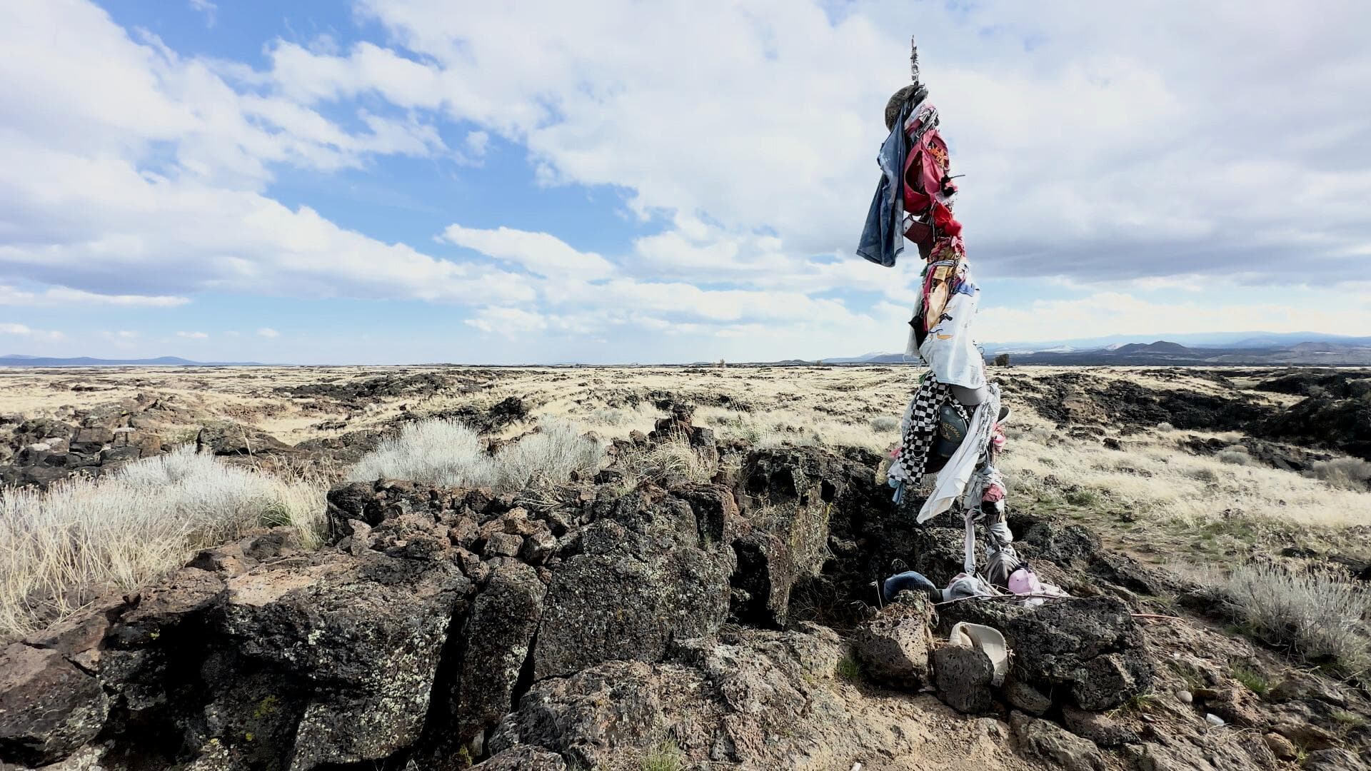A film still from Black Glass shows an open field, presumably atop a mountain, with algae speckled rocks covered in yellow grass. There is a blue sky filled with fluffy white clouds. In the foreground a pole stands planted between the rocks, draped with hats, shirts, and other articles of clothing.
