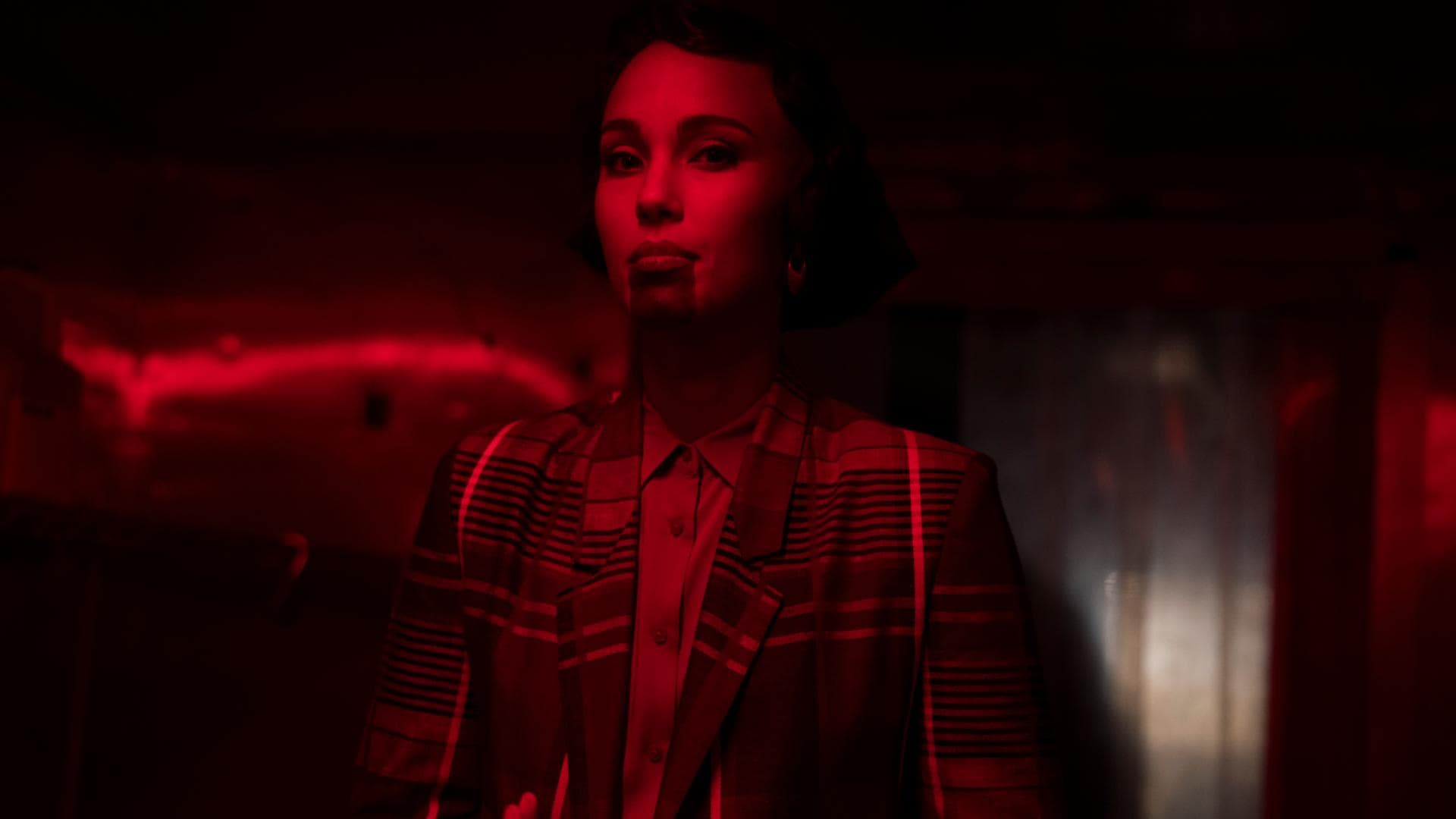 A film still from BLKNWS: Terms & Conditions shows a woman standing in a room illuminated by red lights. There is a thick dark ink that goes from the bottom of her lip to her chin. Her hands are stacked on top of each other palm up in front of her. She is wearing a plaid suit.