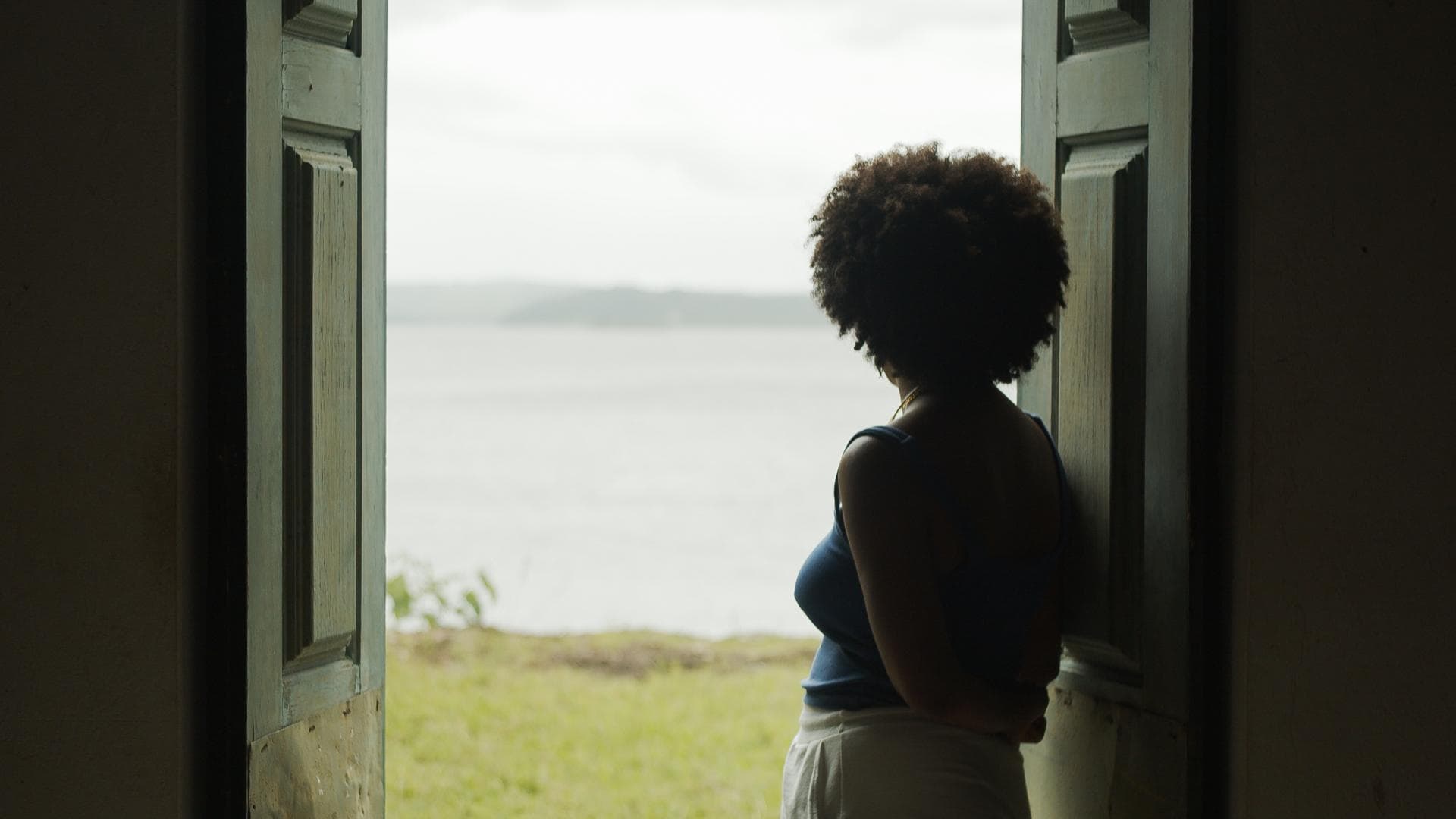 A film still from Cais shows the back of a woman standing at the opening of double doors. She looks out at a stretch of grass that leads up to the grayscale water and sky in front of her.