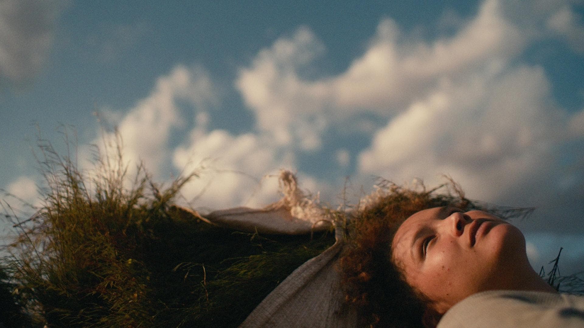 A film still from Carissa shows a young girl lying against white bags filled with a green crop. The girl is looking into the partly cloudy blue sky.