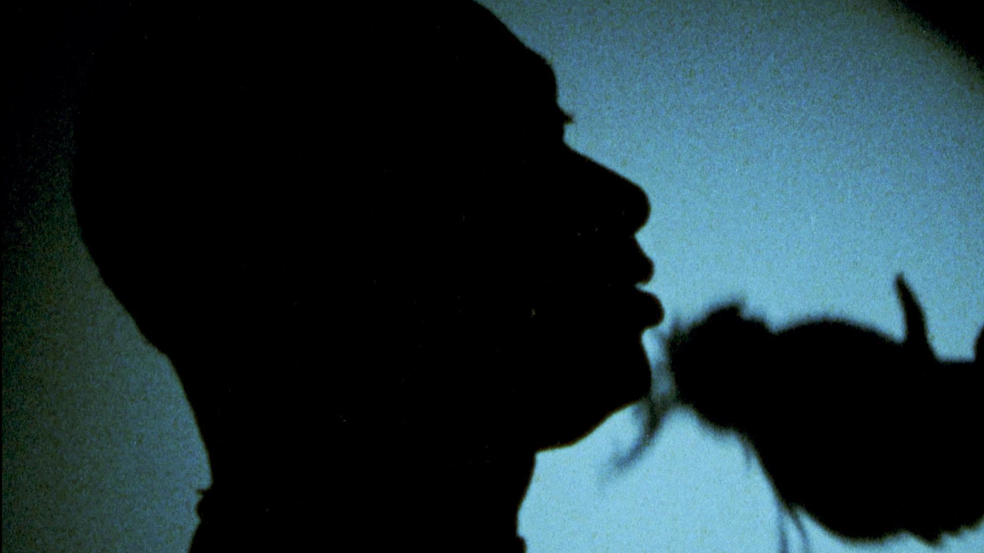 A film still from Dooni shows the silhouette of a person’s head in front of a wall illuminated by an unknown light source. On the wall is the shadow of another head.