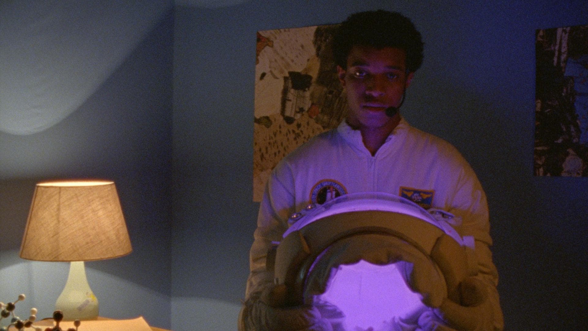 A film still from Highway to the Moon shows a young Black man in an astronaut suit standing in a room with blue walls. He holds his helmet in his hands, which emits a glowing light purple light that softly illuminates his body.