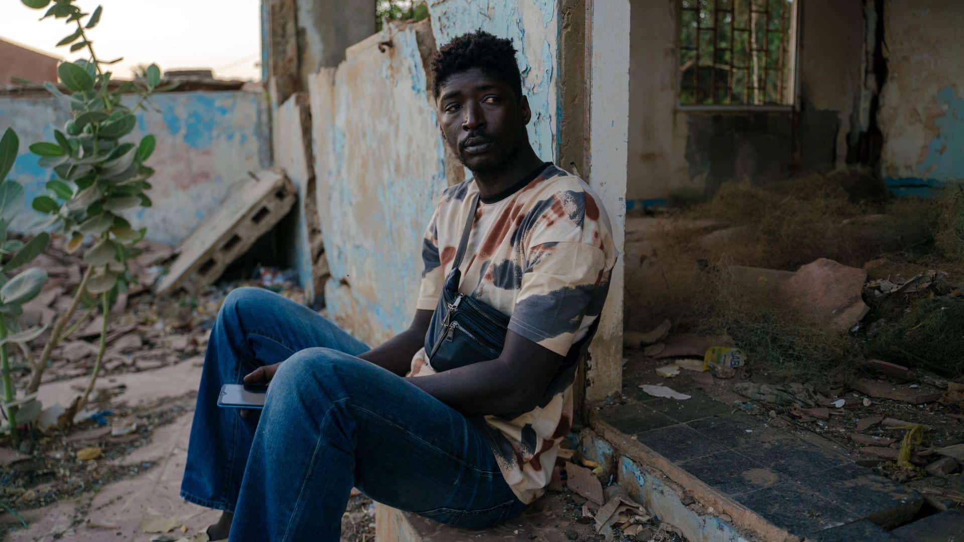 A film still from Lees Waxul shows a Black man sitting on a step with his back against the wall of an abandoned building. The structure is crumbling, with gaps in the walls and plants growing up from the floor. He looks off to the side at something off screen, holding a phone between his legs.