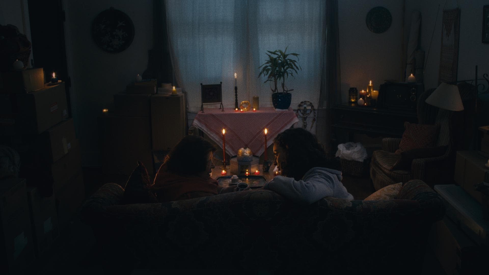 A film still from Maqluba shows a serene living room lit up solely with various candles. From the back of the couch an older woman can be seen speaking to a young woman as they share a close moment.