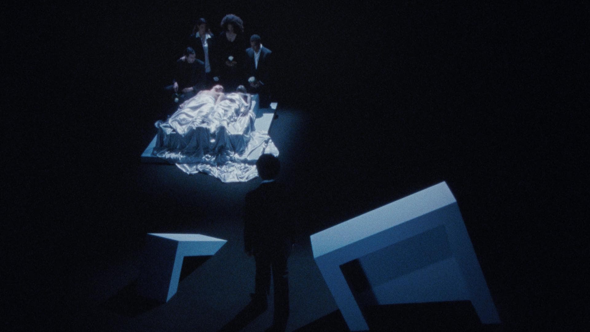 A film still from Oceania shows a dark, seemingly boundless room. A spotlight illuminates a platform holding two bodies, lying face up, covered from the chest down with a sheet. At the head of the platform stands a small group of people dressed in black suits and dresses, each holding a flower. A short distance away, another man in a suit faces them. He stands between two hollowed three-dimensional rectangles that sink into the ground.