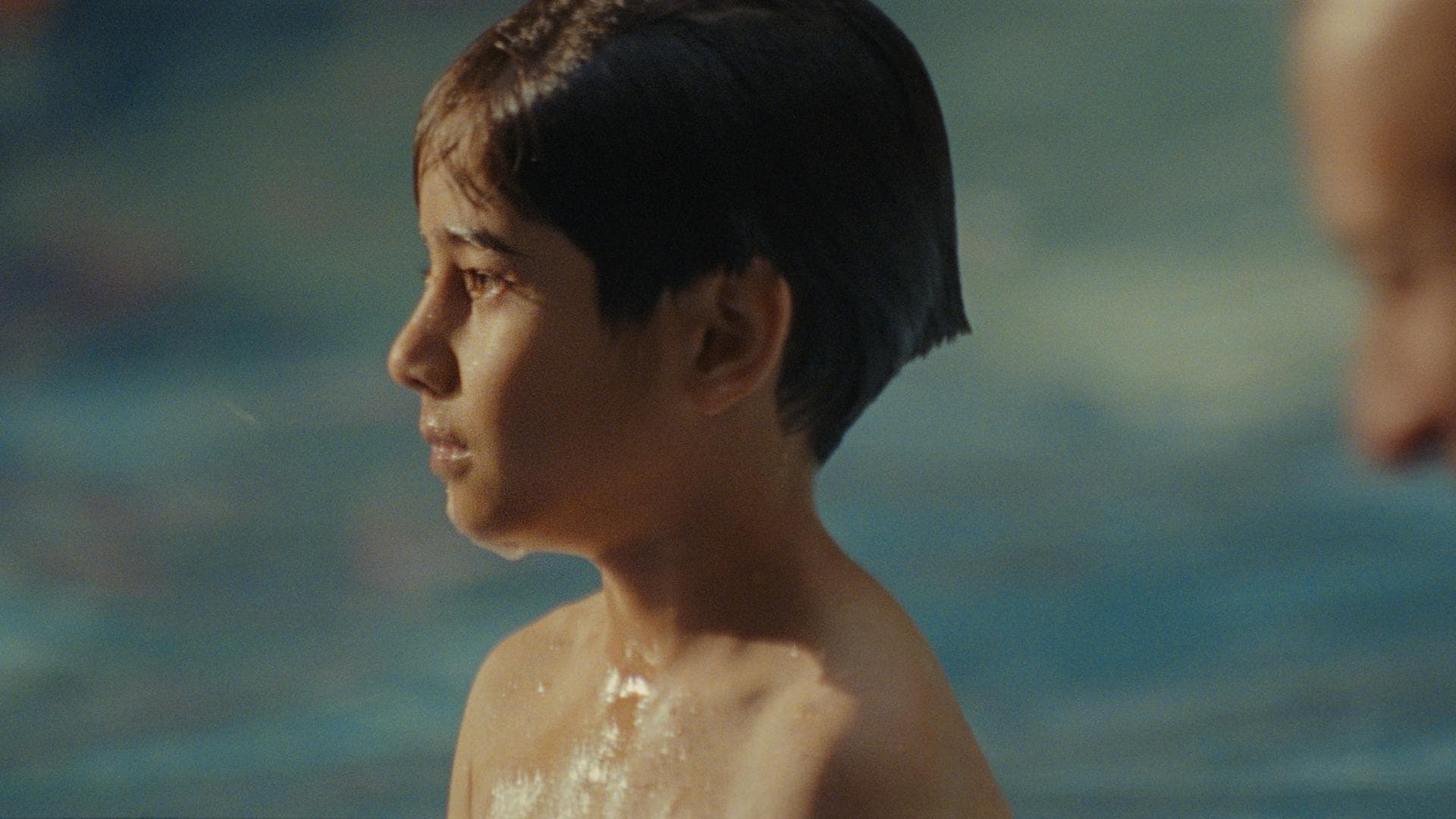 A film still from One Day This Kid shows a young boy standing in a body of water. He looks off into the distance at something off screen with an emotionless face. Everything around the boy is out of focus, including the side profile of a man who stands behind him.