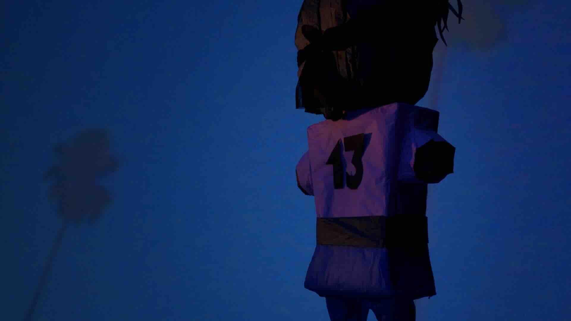 A film still from Piñata Prayers shows the back of a piñata that faces a blue wall with a palm tree pattern. The piñata resembles a person playing a sport. It wears the number thirteen on its back.