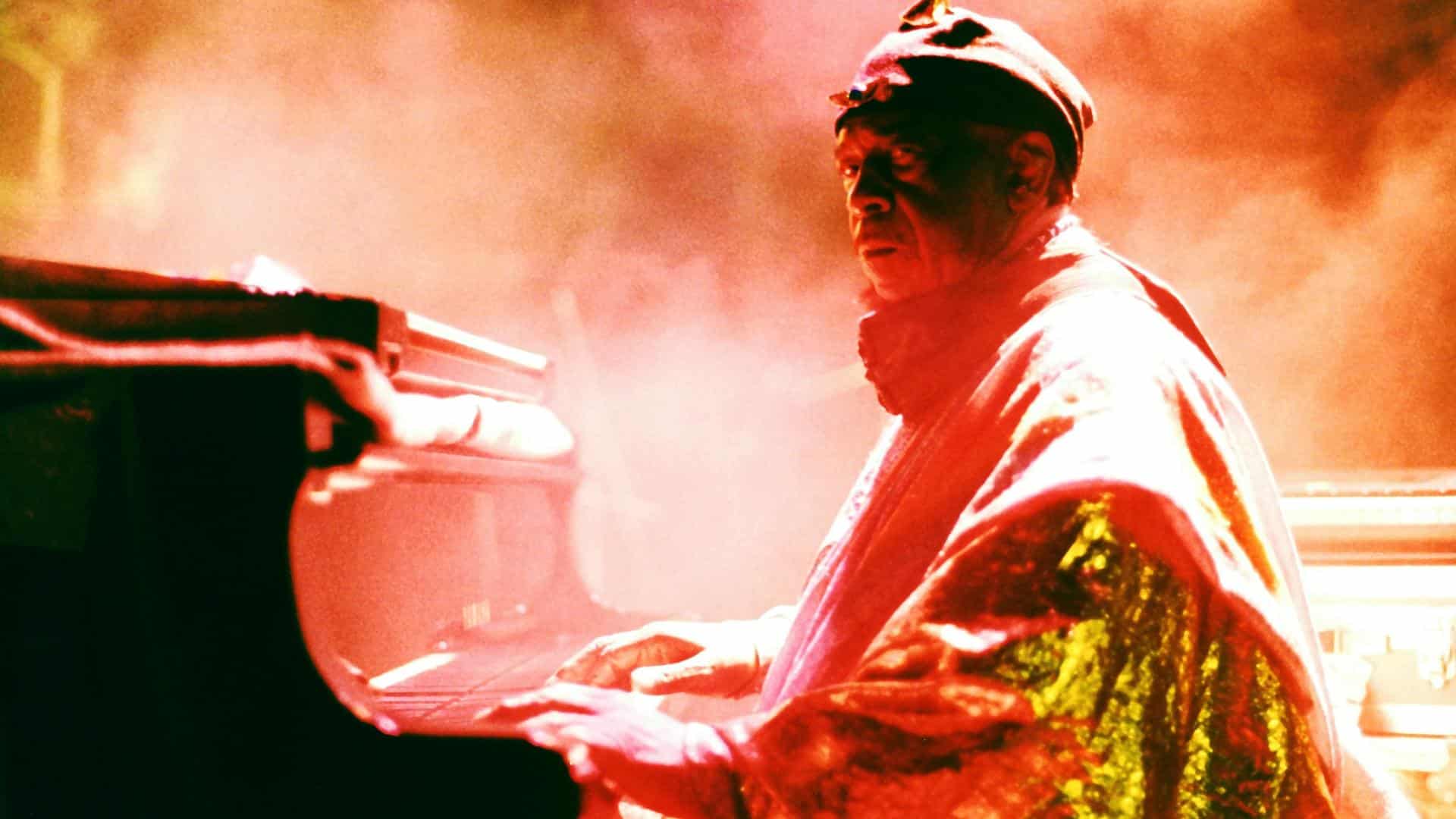 A film still from Sun Ra: Do the Impossible shows a Black man dressed in a red and metallic gold outfit seated at a piano. He looks to the side, into the camera. A warm colored fog surrounds him, filling the space with a hazy glow.