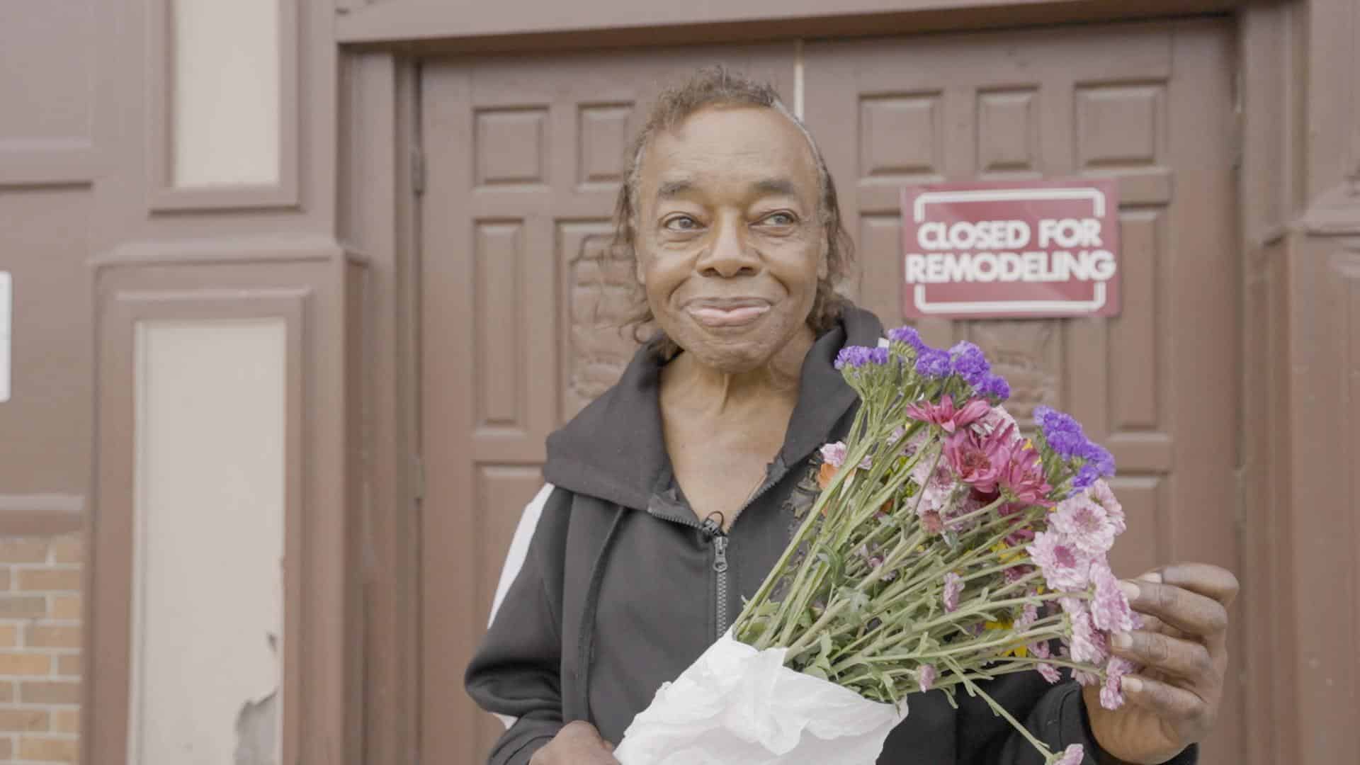 A film still from Taking Walls shows an older Black woman standing at the doors of a brown building. A small sign on one of the doors reads: “Closed for Remodeling”. The woman grins, holding a bouquet of purple and pink flowers, and looks to the side off into the distance.