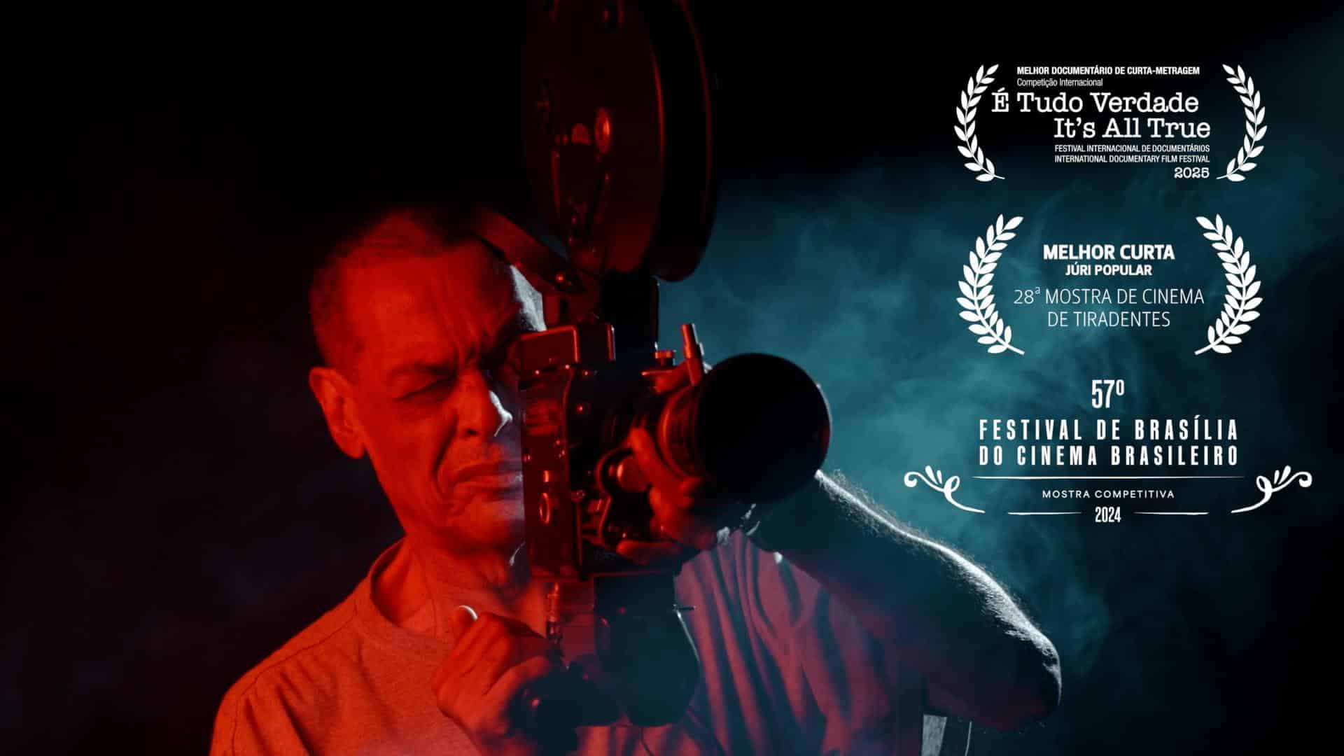 A film still from The Nile Shows a man in red lighting holding a film camera up to his eye. A light fog is casted from a spotlight on his side.